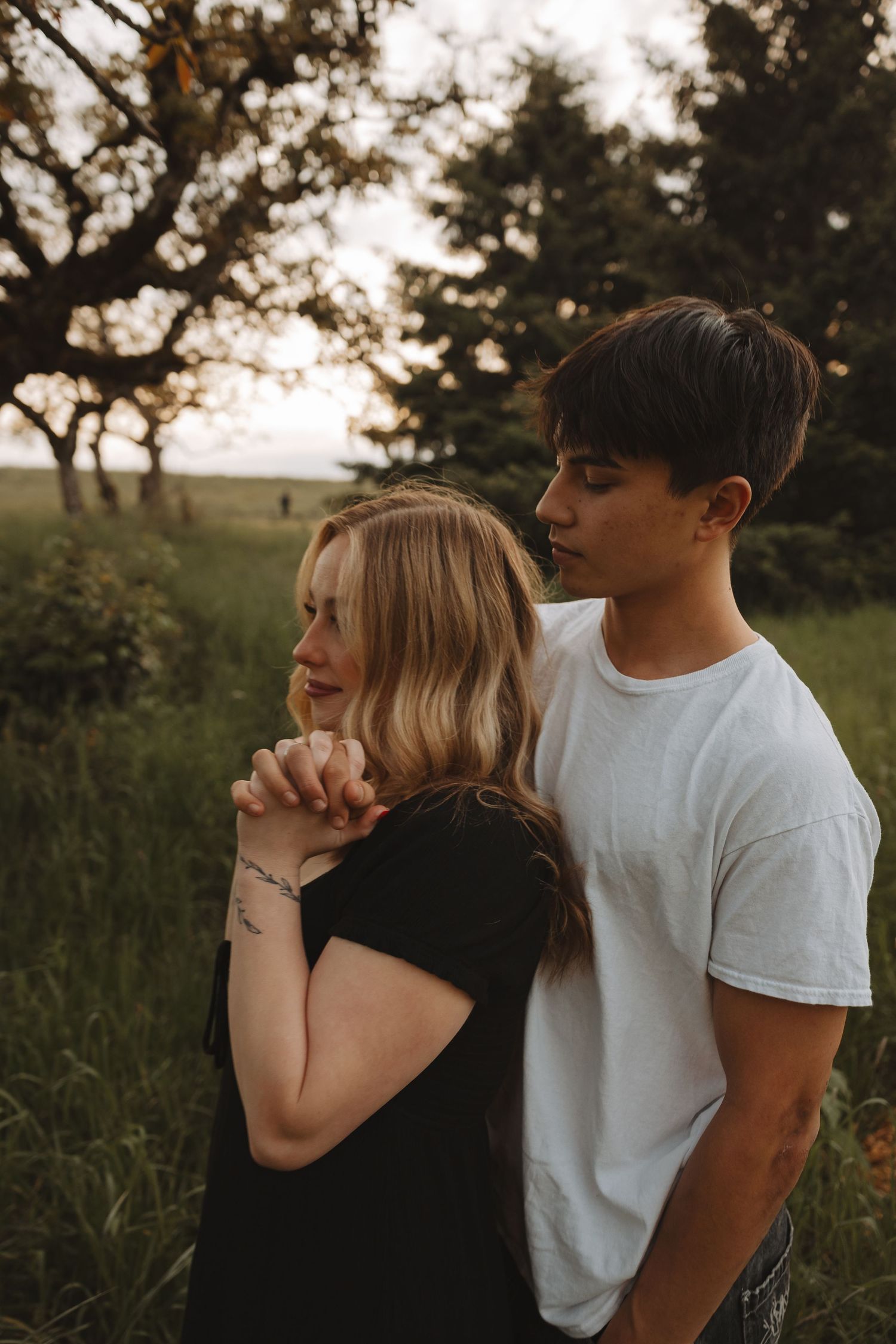 A young couple embraces in a lush green field at sunset, the man in white holding the woman in black close.
