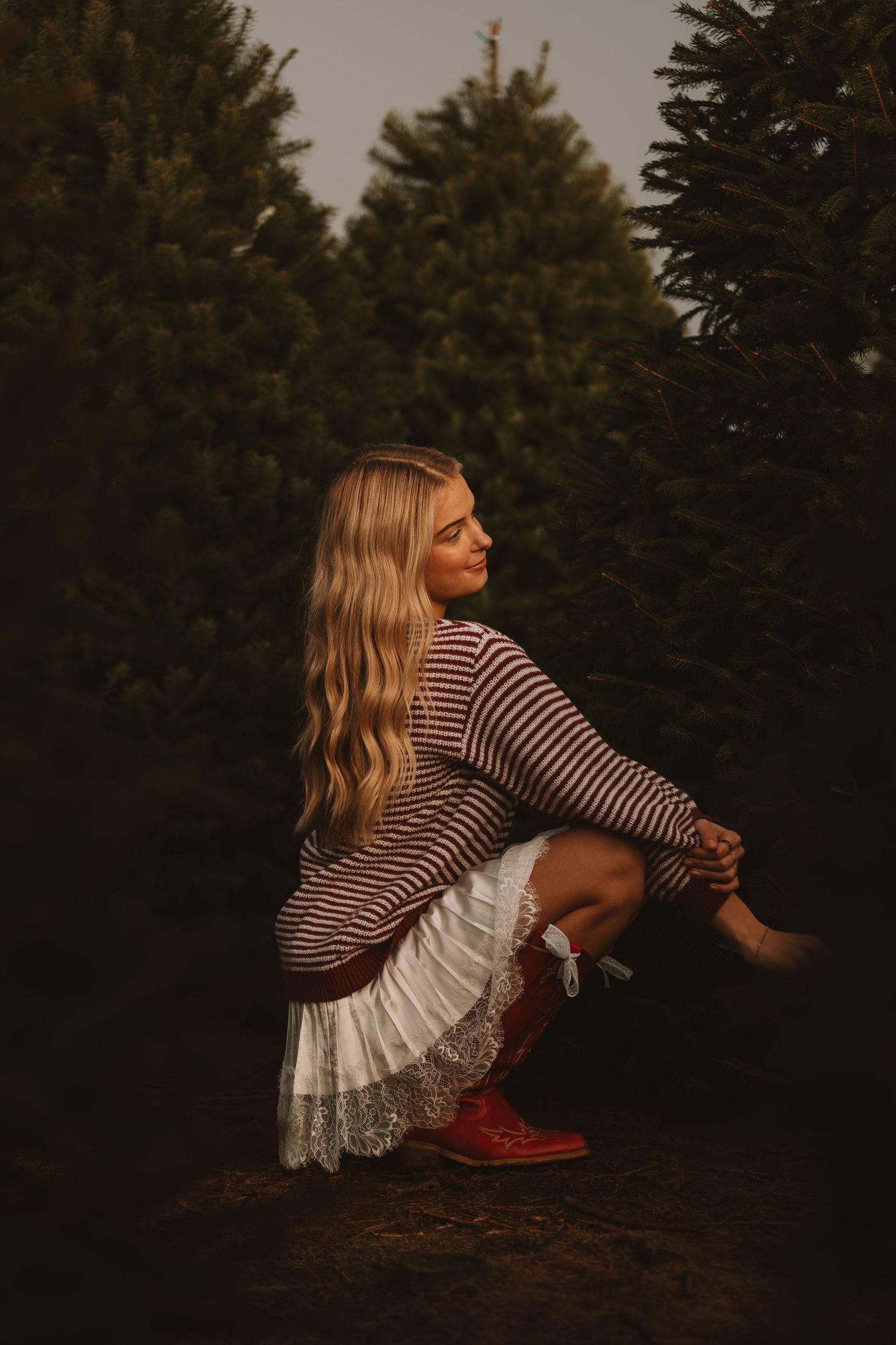 Person in striped shirt and white skirt sits casually against evergreen trees at dusk.