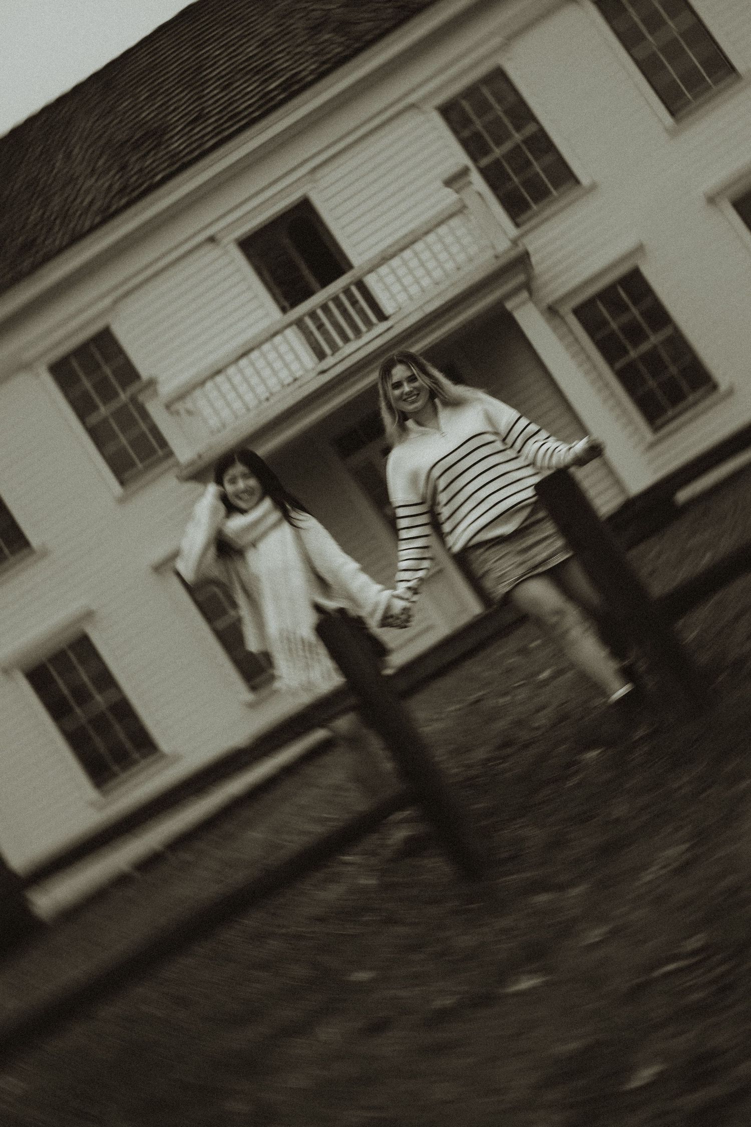 Black and white photo showing two people holding hands and running in front of a white colonial-style house.