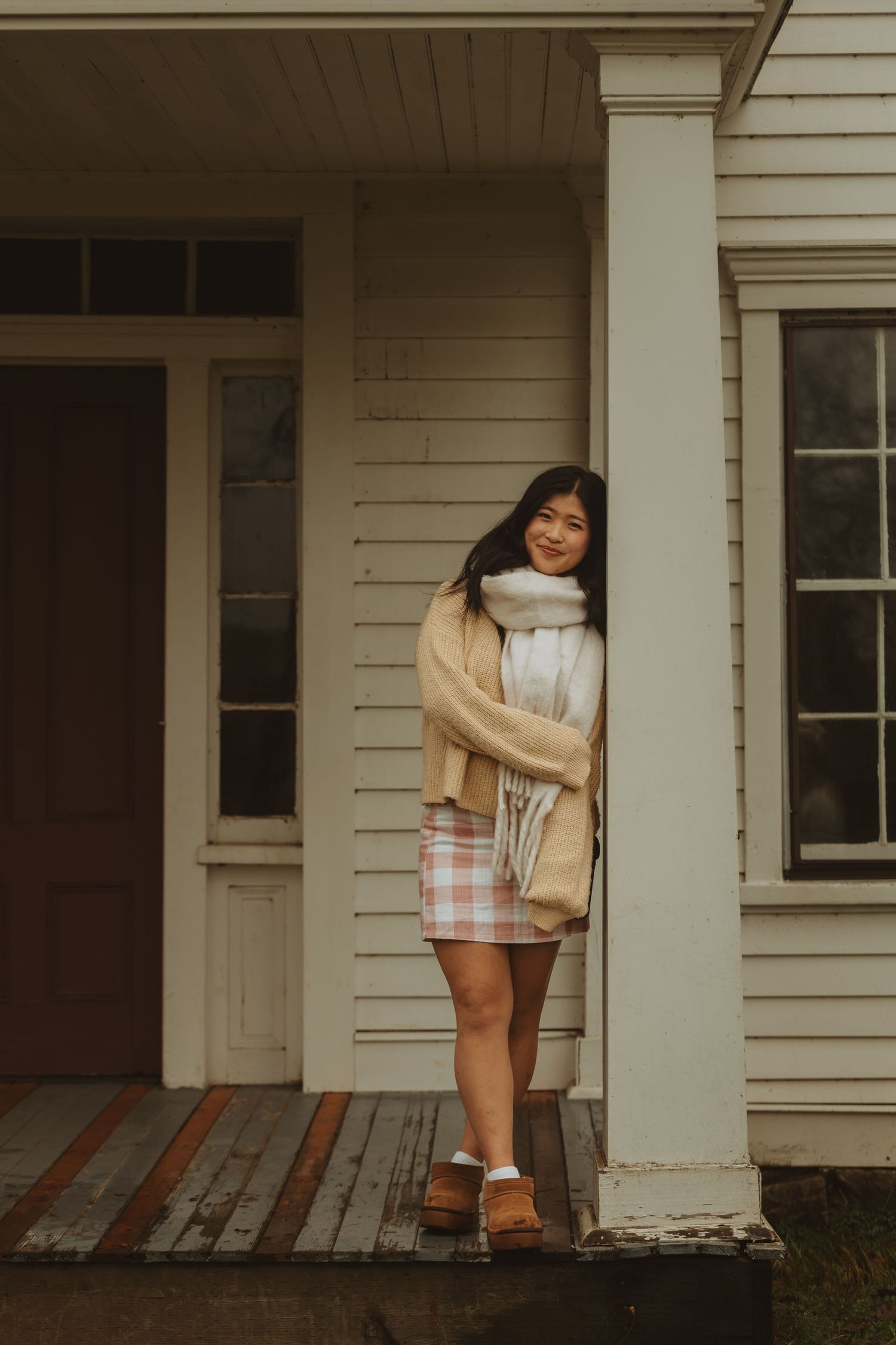 Person in beige sweater and plaid skirt posing on white farmhouse porch.