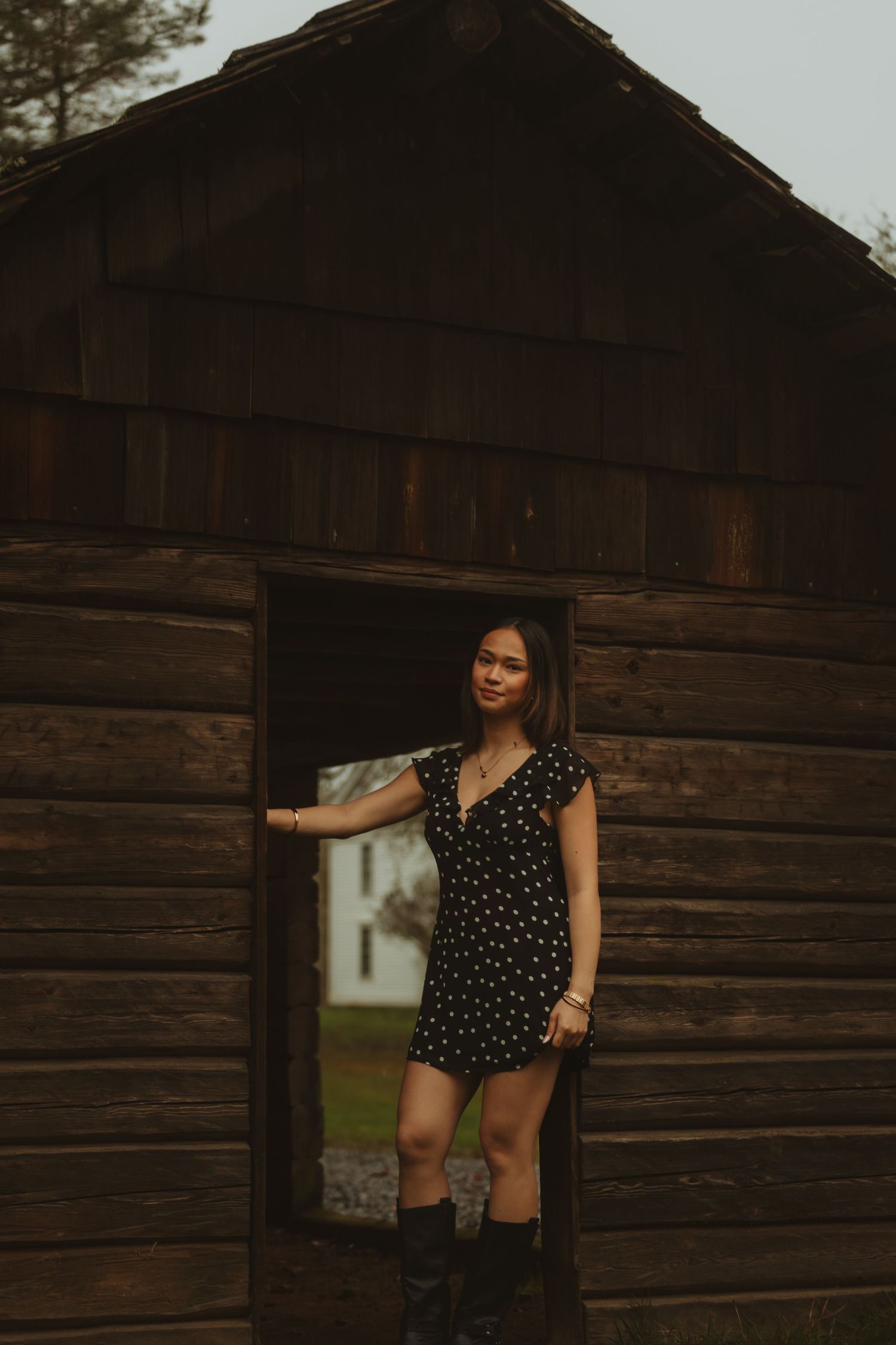 Woman in polka dot dress poses against rustic wooden barn wall in moody evening lighting.