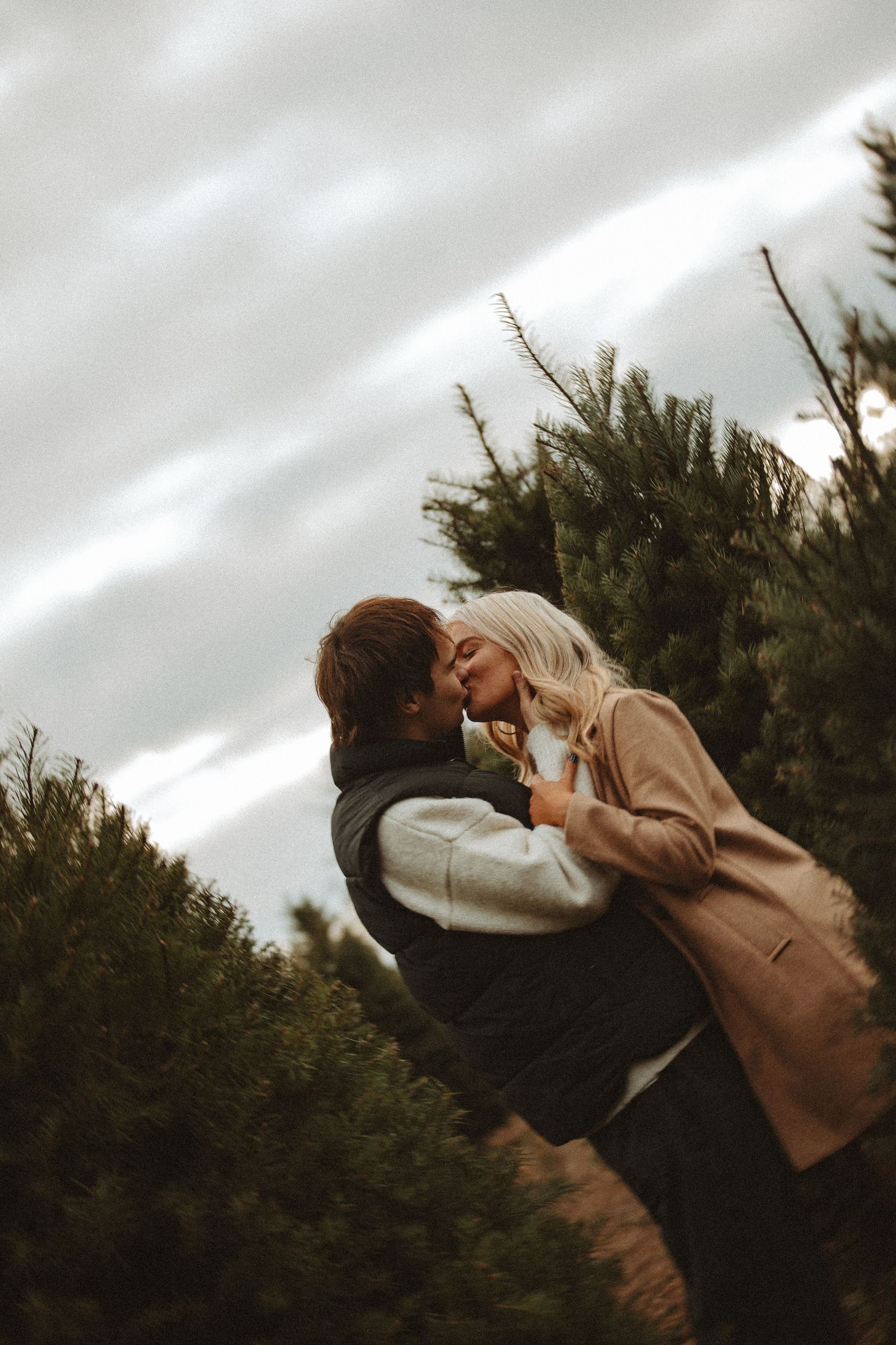 A romantic couple shares a tender kiss against a backdrop of evergreen trees and moody sky.
