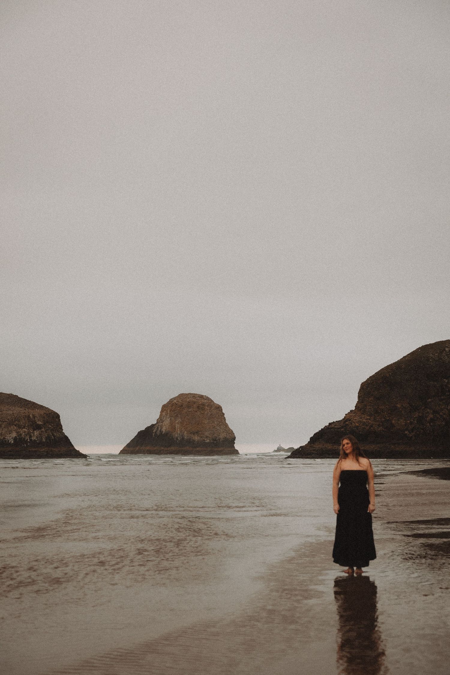 A dramatic coastal landscape with rock formations and a lone figure on a foggy beach during low tide.