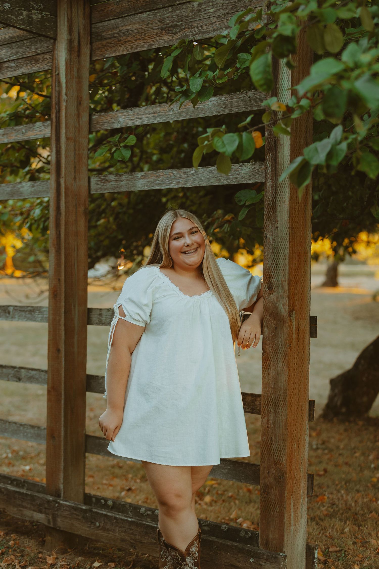 A person in a white dress poses casually against a rustic wooden fence during golden hour.