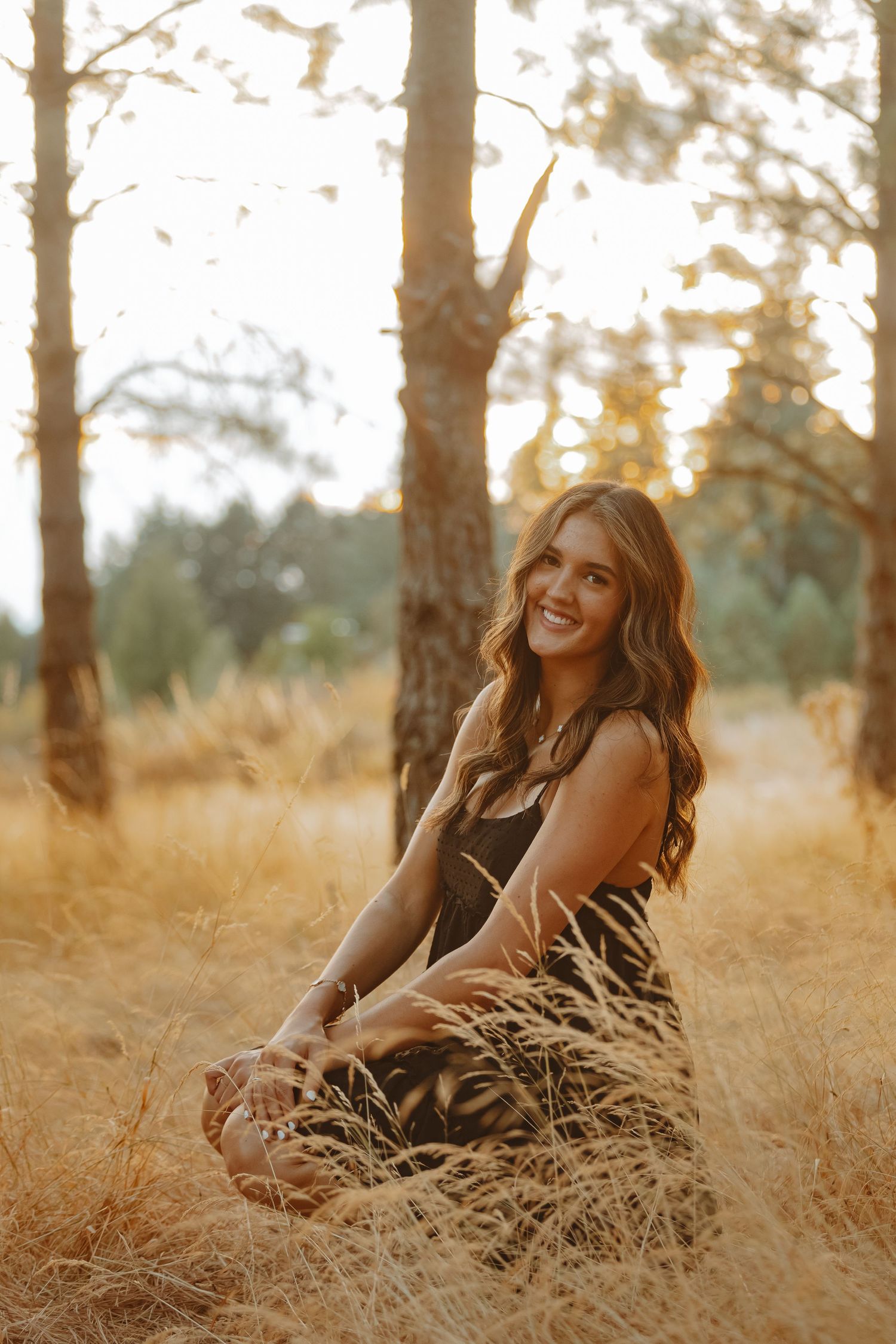 A person sits in golden grass at sunset among tall trees in a dreamy autumn setting.