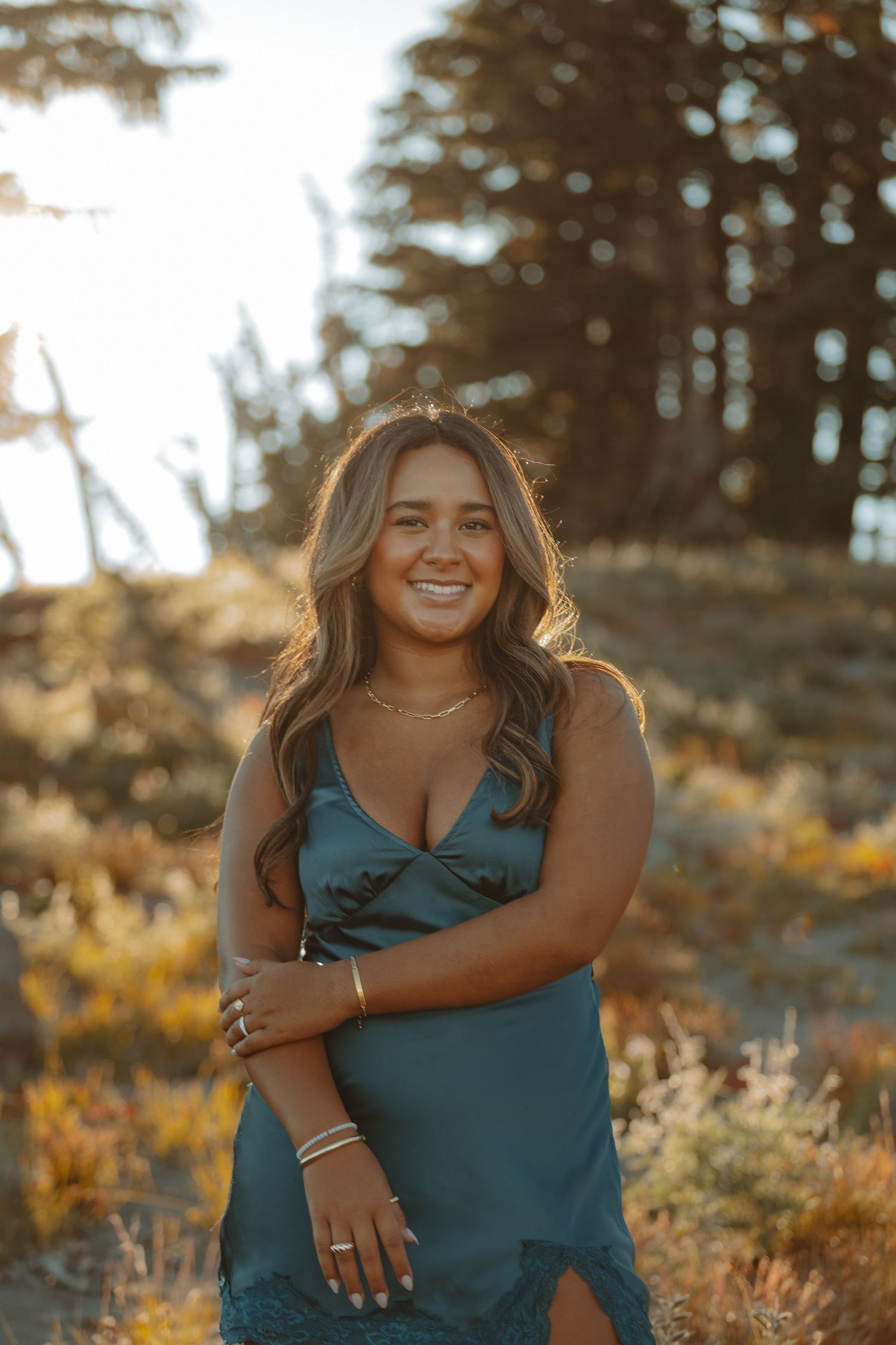 A woman in a teal dress poses outdoors during golden hour with trees and sunlight in the background.