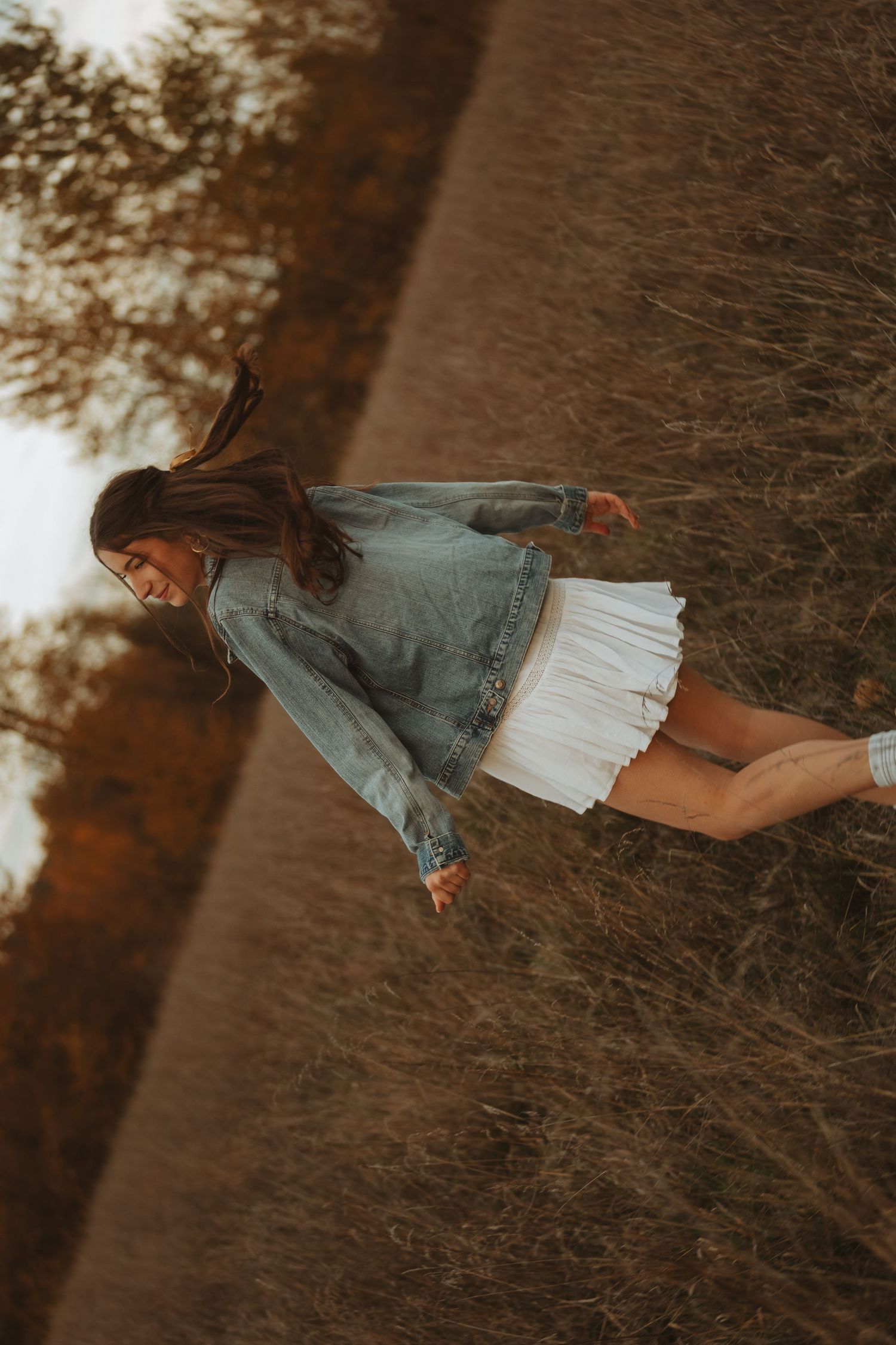 Person in denim and white dress twirling in a field during sunset.
