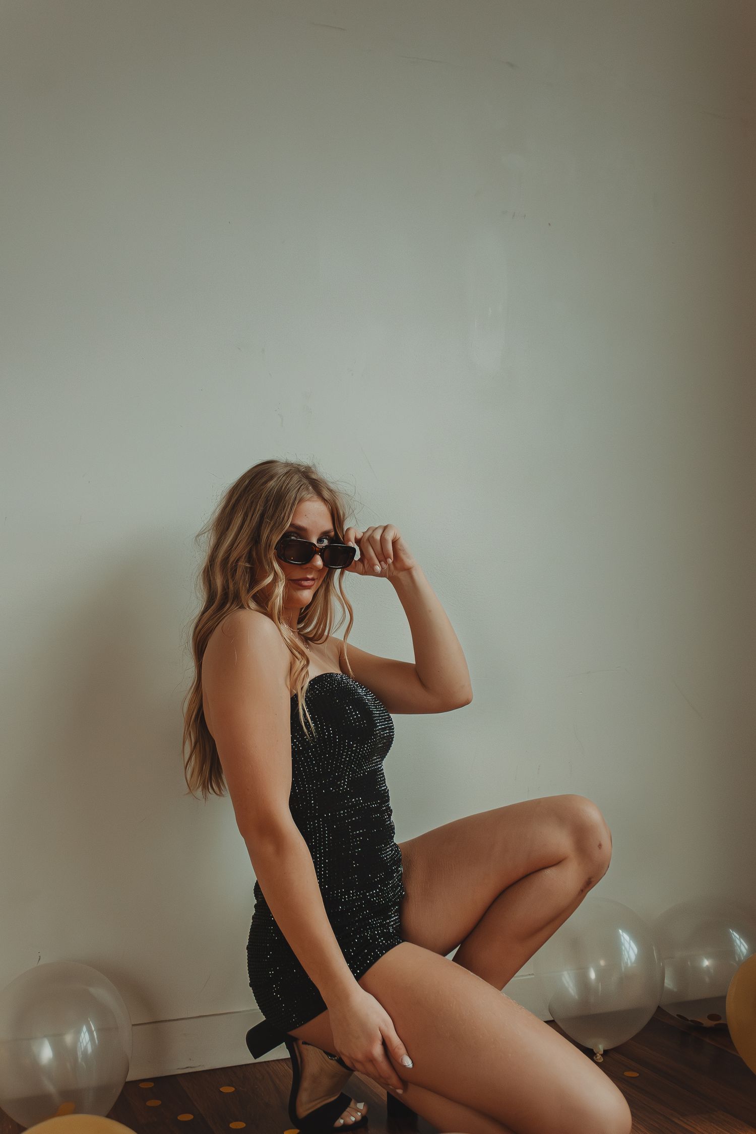Person in black dress and sunglasses posing with balloons on hardwood floor.