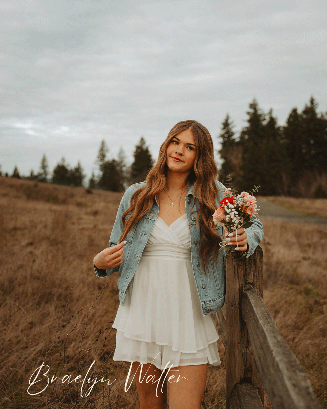 Person wearing white dress and denim jacket poses with flowers in a countryside setting.