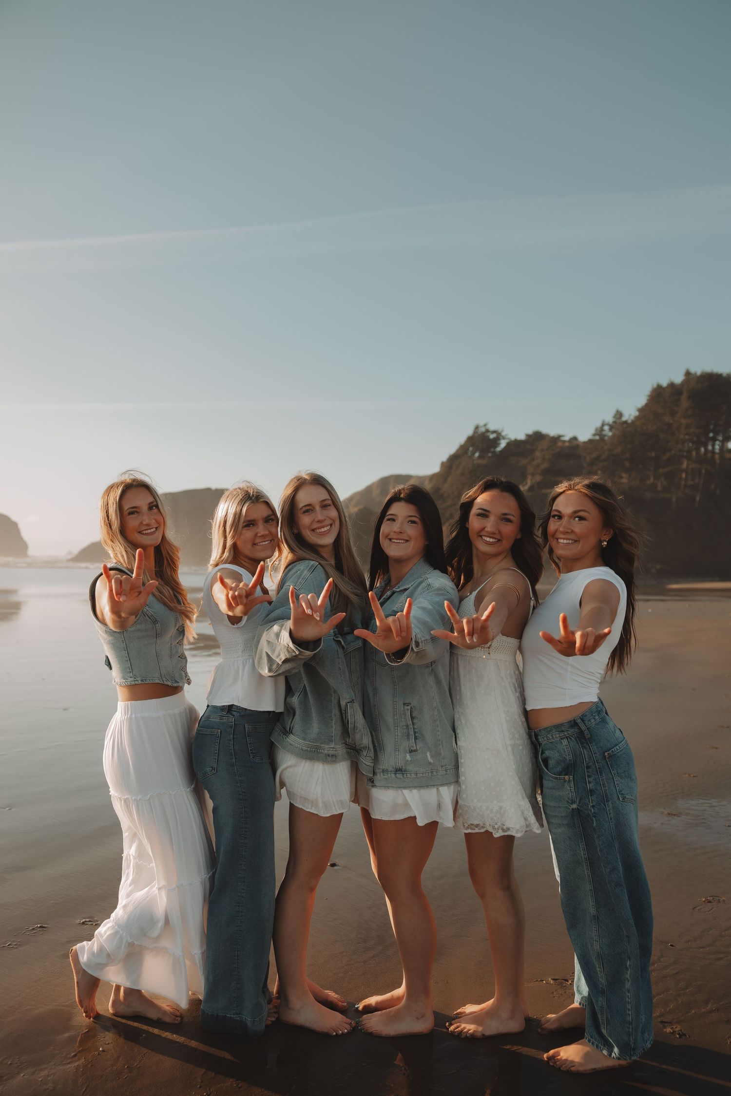 Six smiling women flash peace signs on a beach at sunset in white and denim outfits.