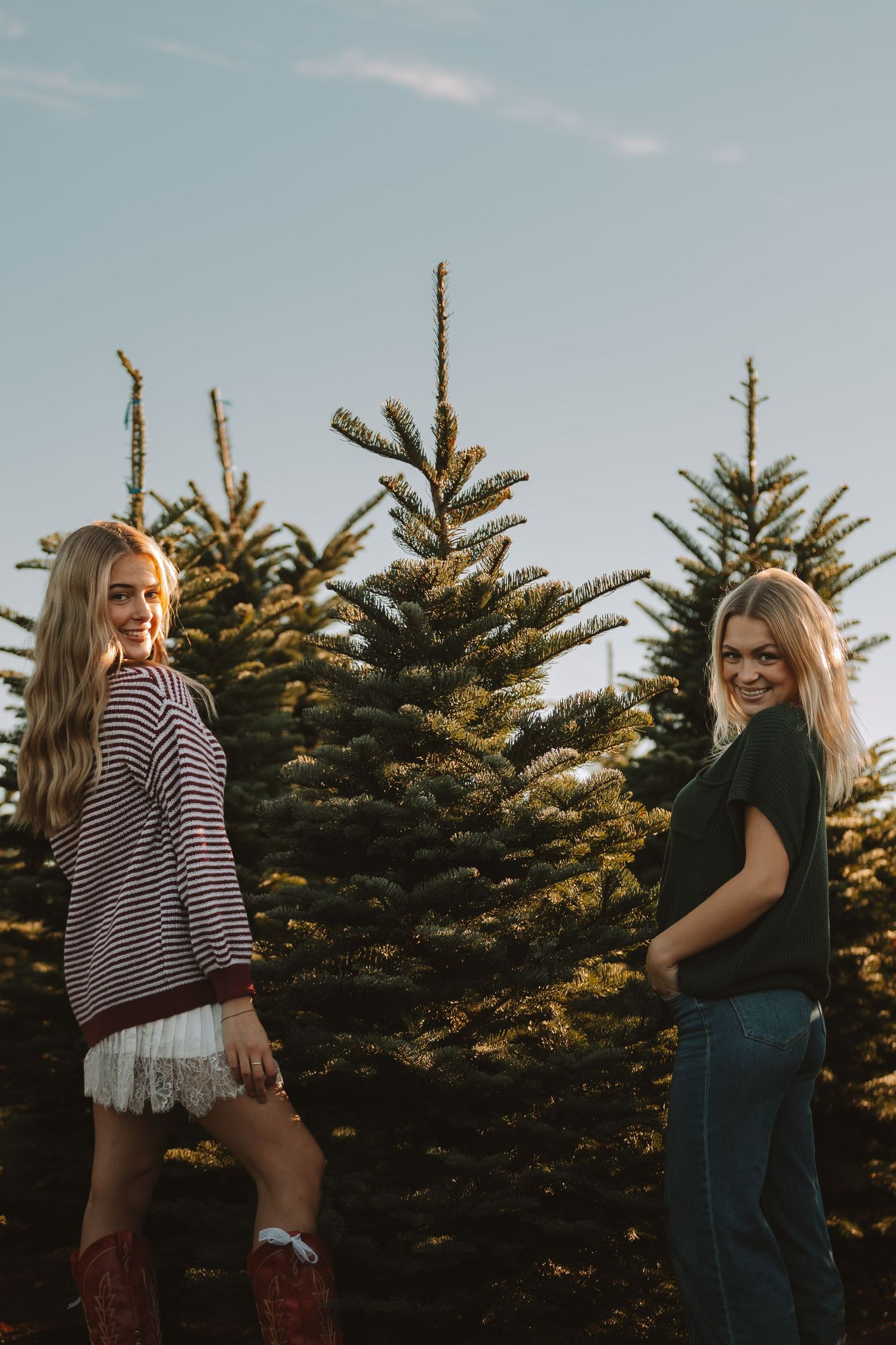 Two people in casual outfits pose together in Christmas tree farm at sunset.