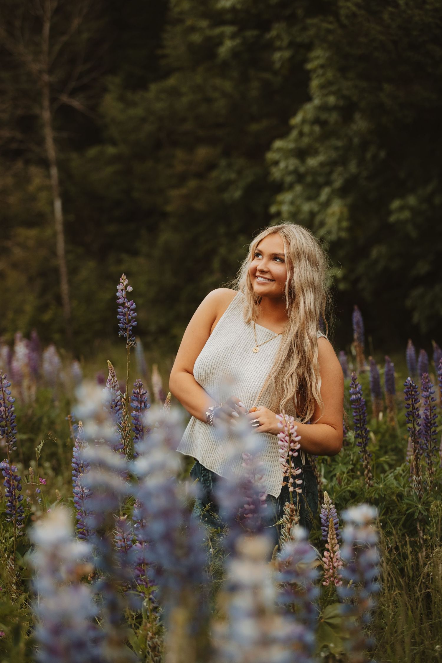 Woman in white tank top standing among blooming purple lupine flowers.