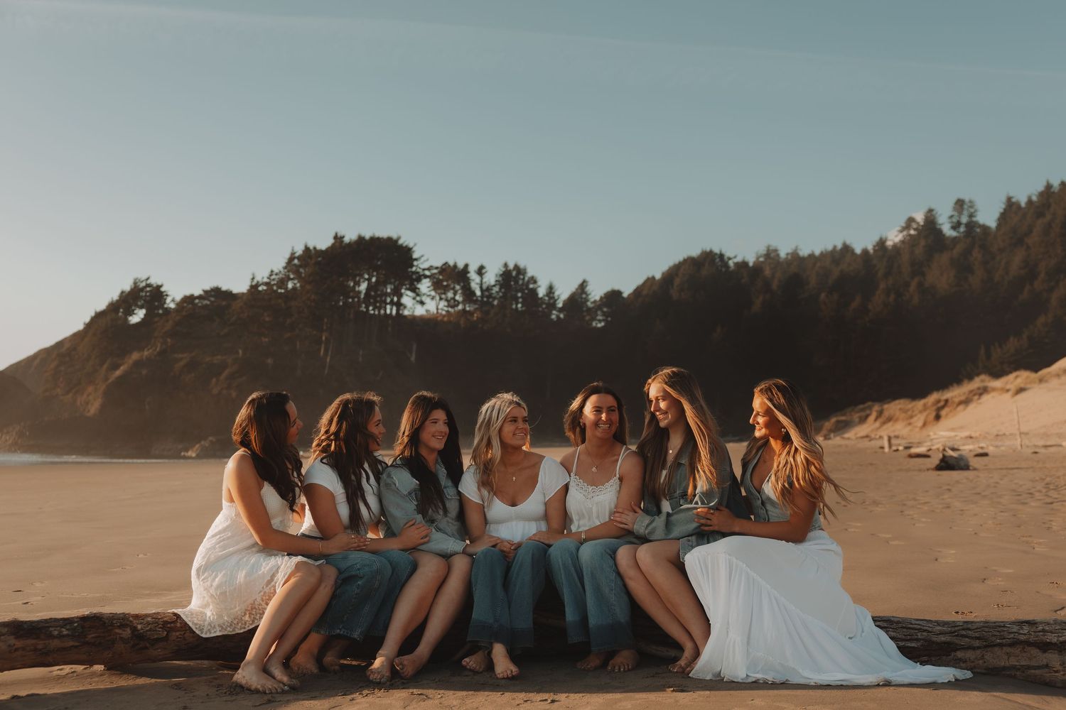 Bride and bridesmaids in white and blue dresses sitting together on driftwood at a scenic beach during golden hour.
