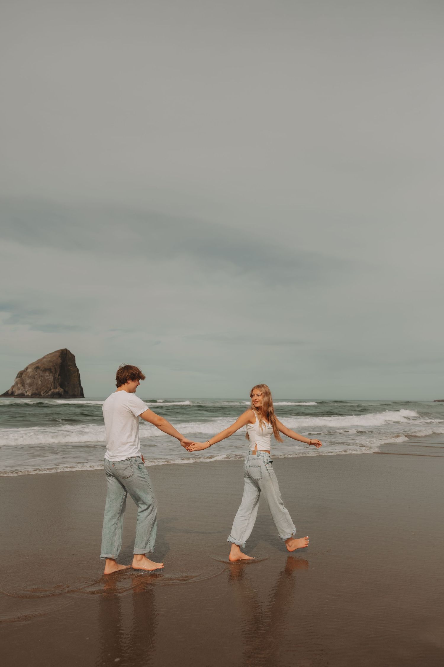 Couple holds hands walking barefoot on wet beach sand with sea stack in background.