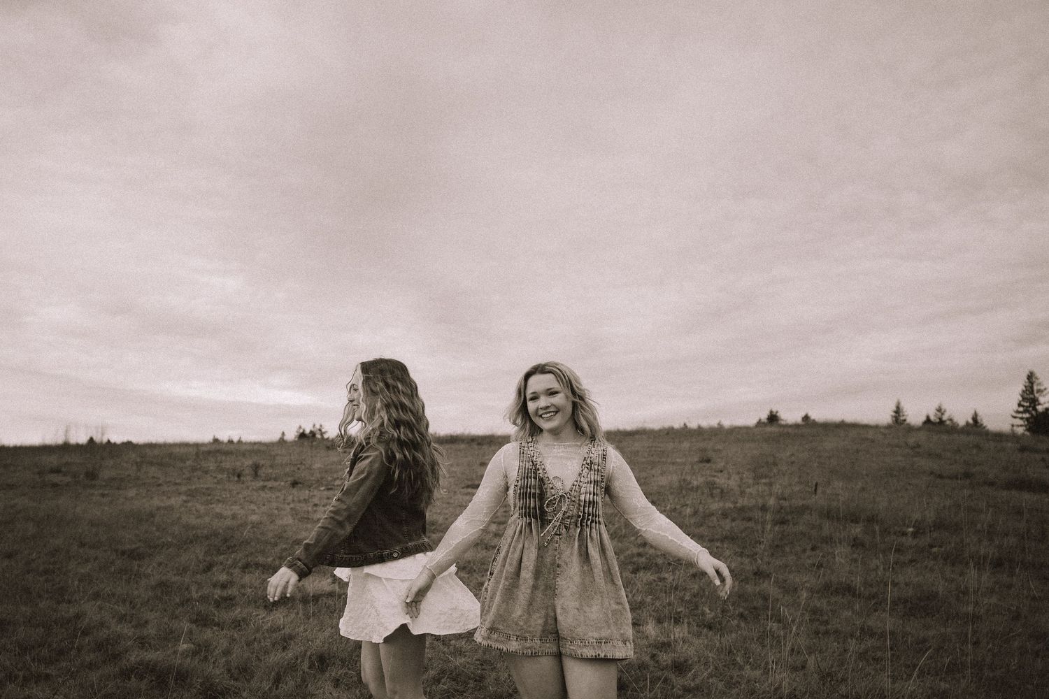 Two people in flowing dresses dance in a field under overcast skies in black and white.