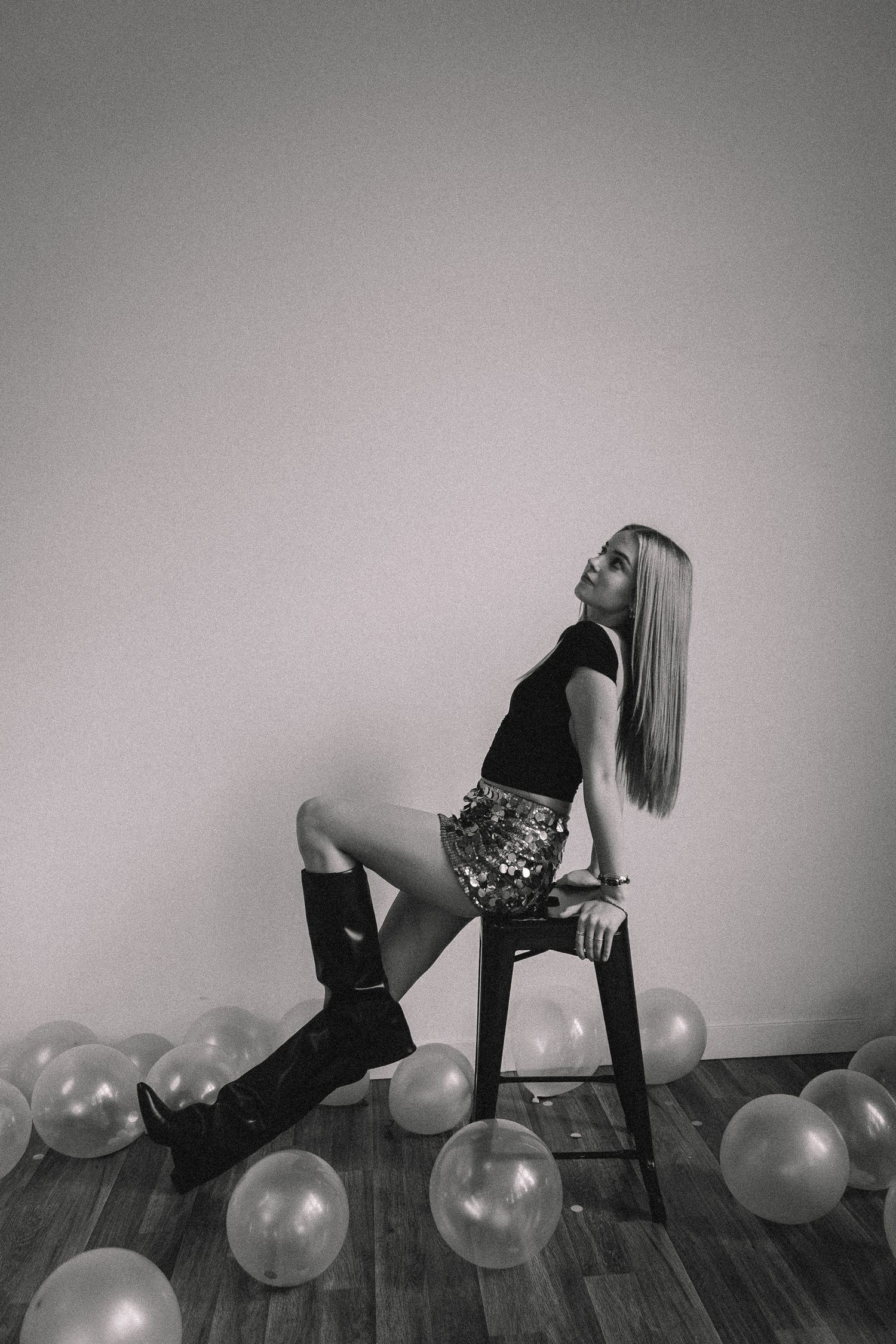 Black and white artistic portrait with balloons on floor featuring dramatic pose on tall stool.