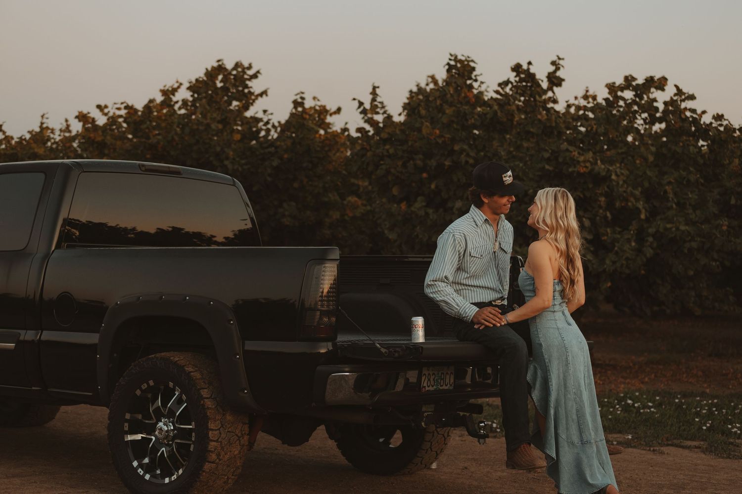 Couple leans against black truck at dusk near lush orchard.