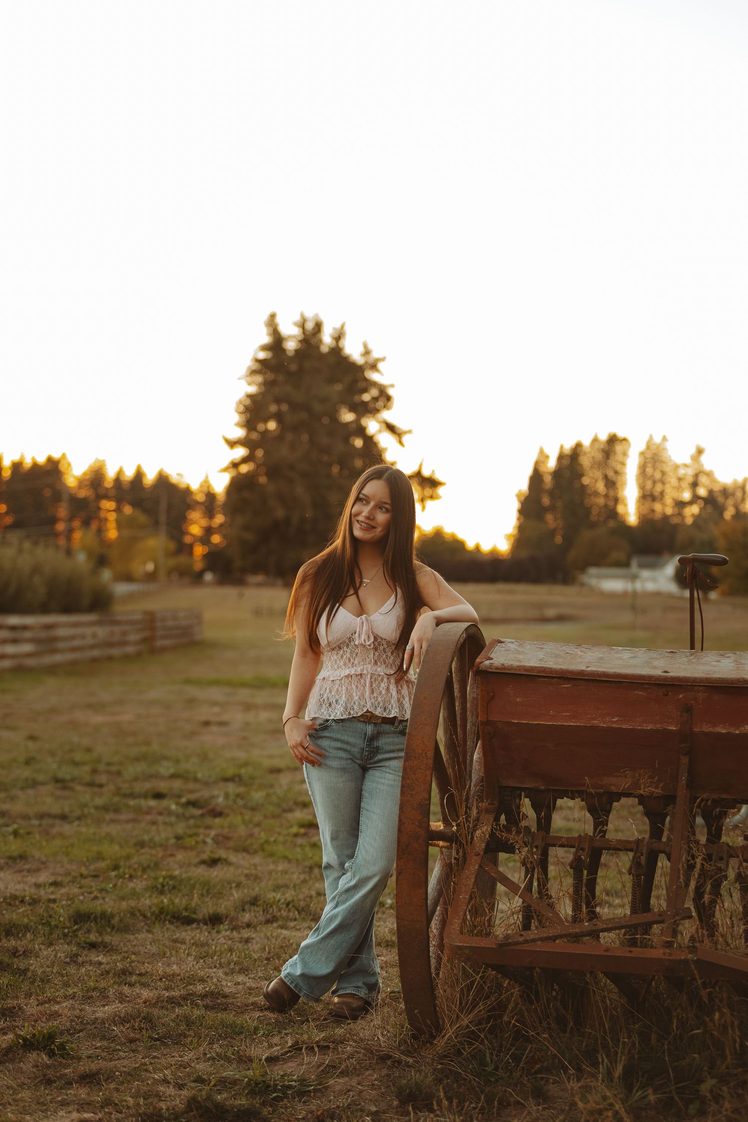 Young woman in lace top and jeans leaning against a rustic wooden wagon at golden hour on a farm.