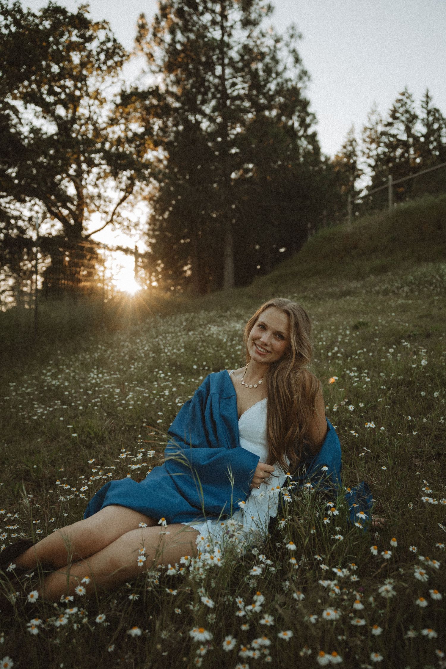 Smiling woman in blue wrap sitting in wildflower meadow with sunset through trees behind her.