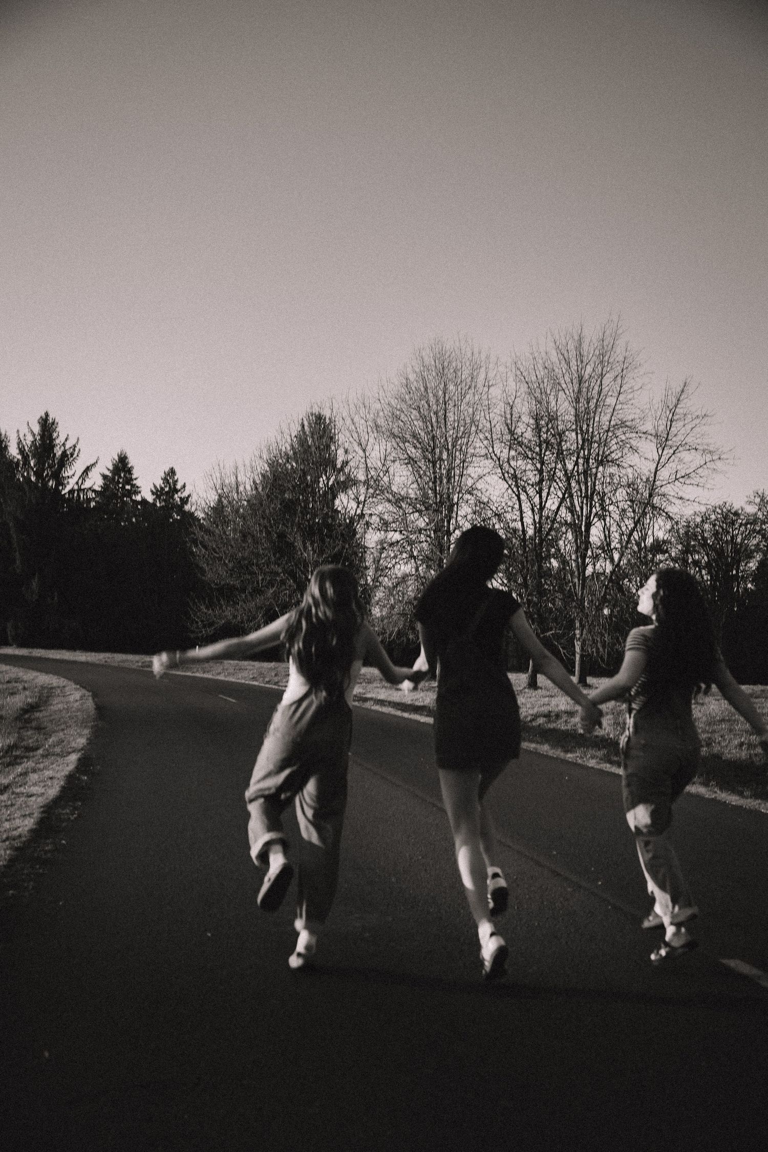 Three girls holding hands walking down a road surrounded by trees in black and white.