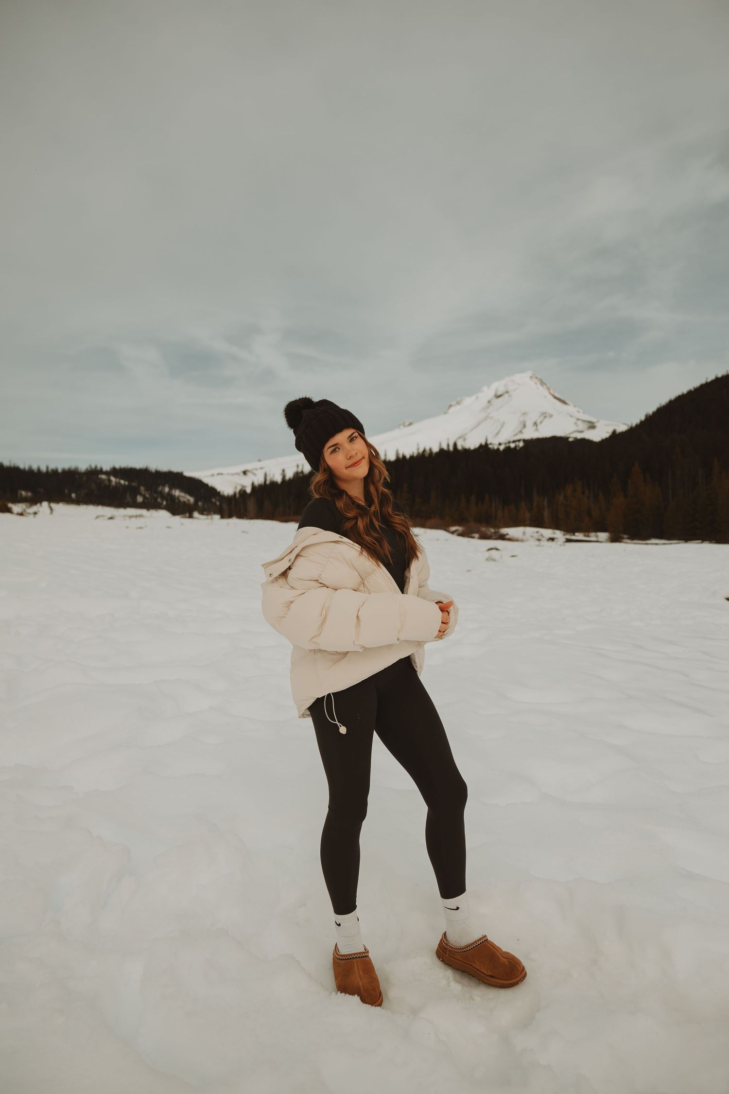 Young woman in cream puffer jacket black leggings and UGG boots poses in snowy mountain landscape.