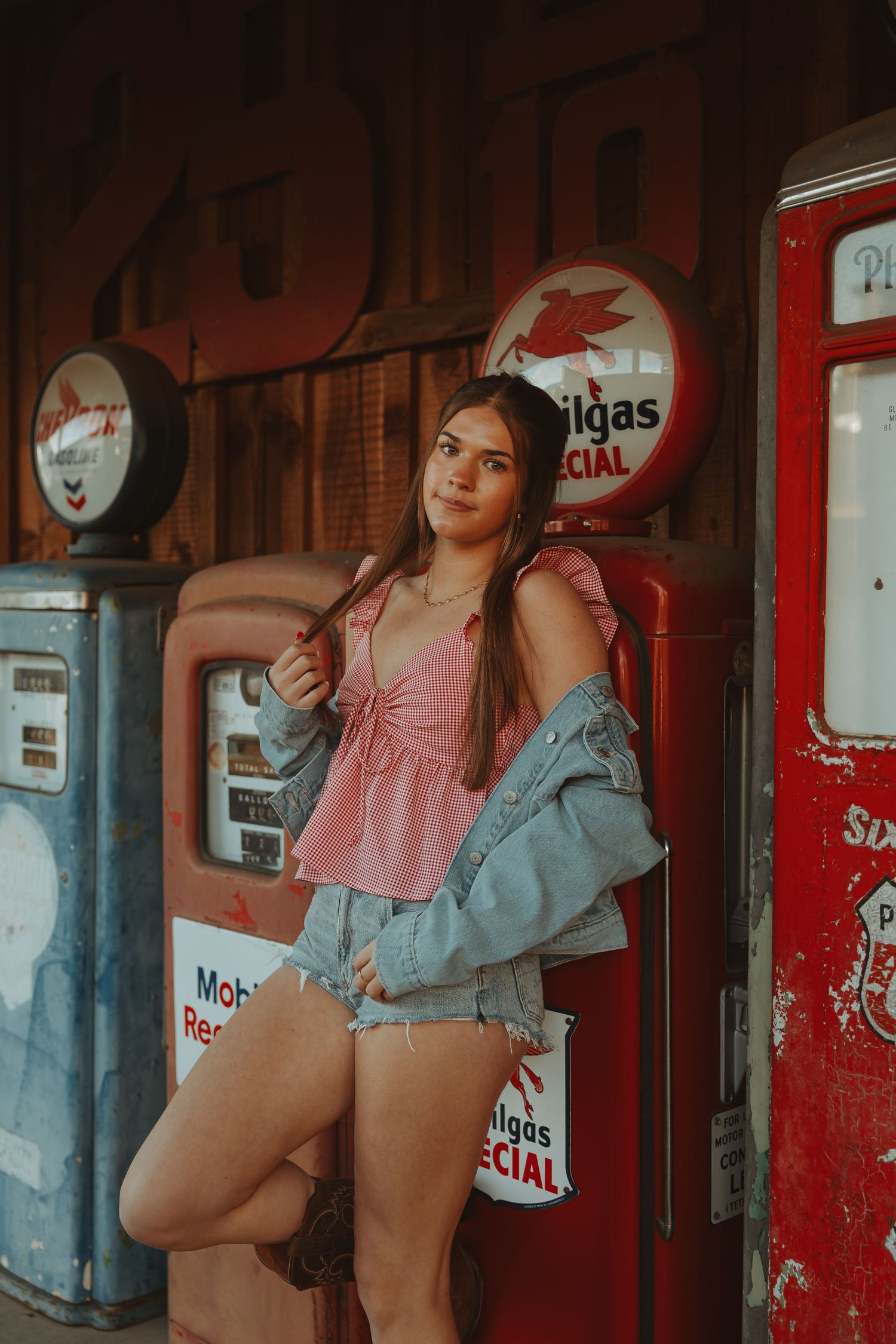 Young woman in pink top, denim jacket, and shorts poses by vintage Mobilgas Special gas pumps at retro station.