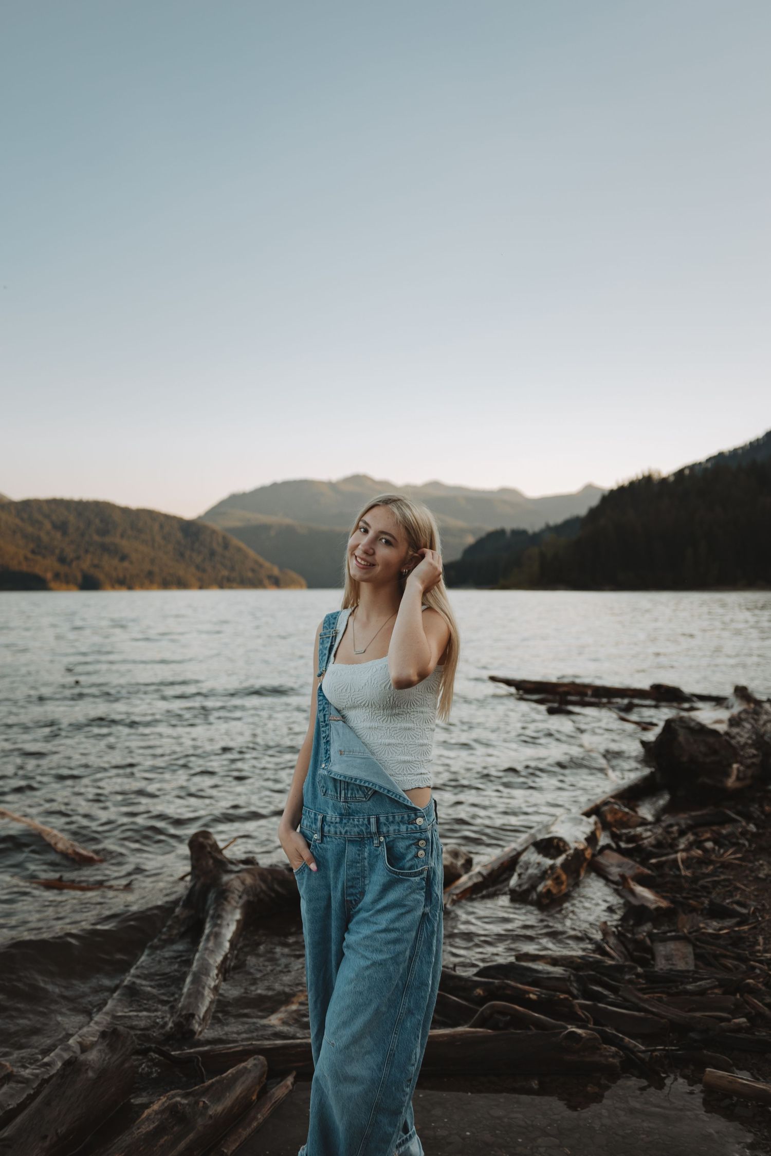 Blonde woman in denim overalls posing on driftwood logs by a scenic mountain lake at sunset.