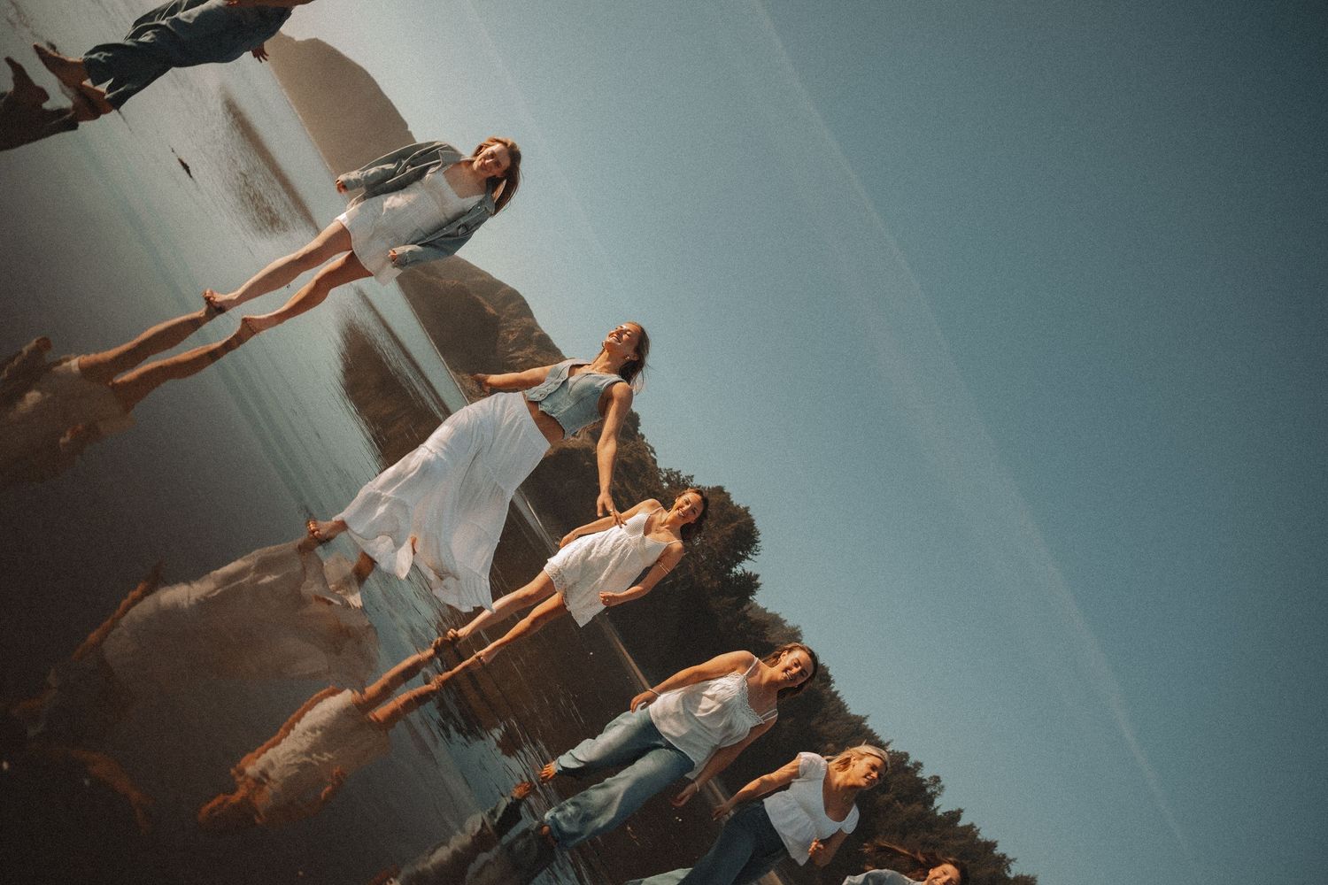 Group of women in white dresses posing by a reflective lake in a creative rotated photo.