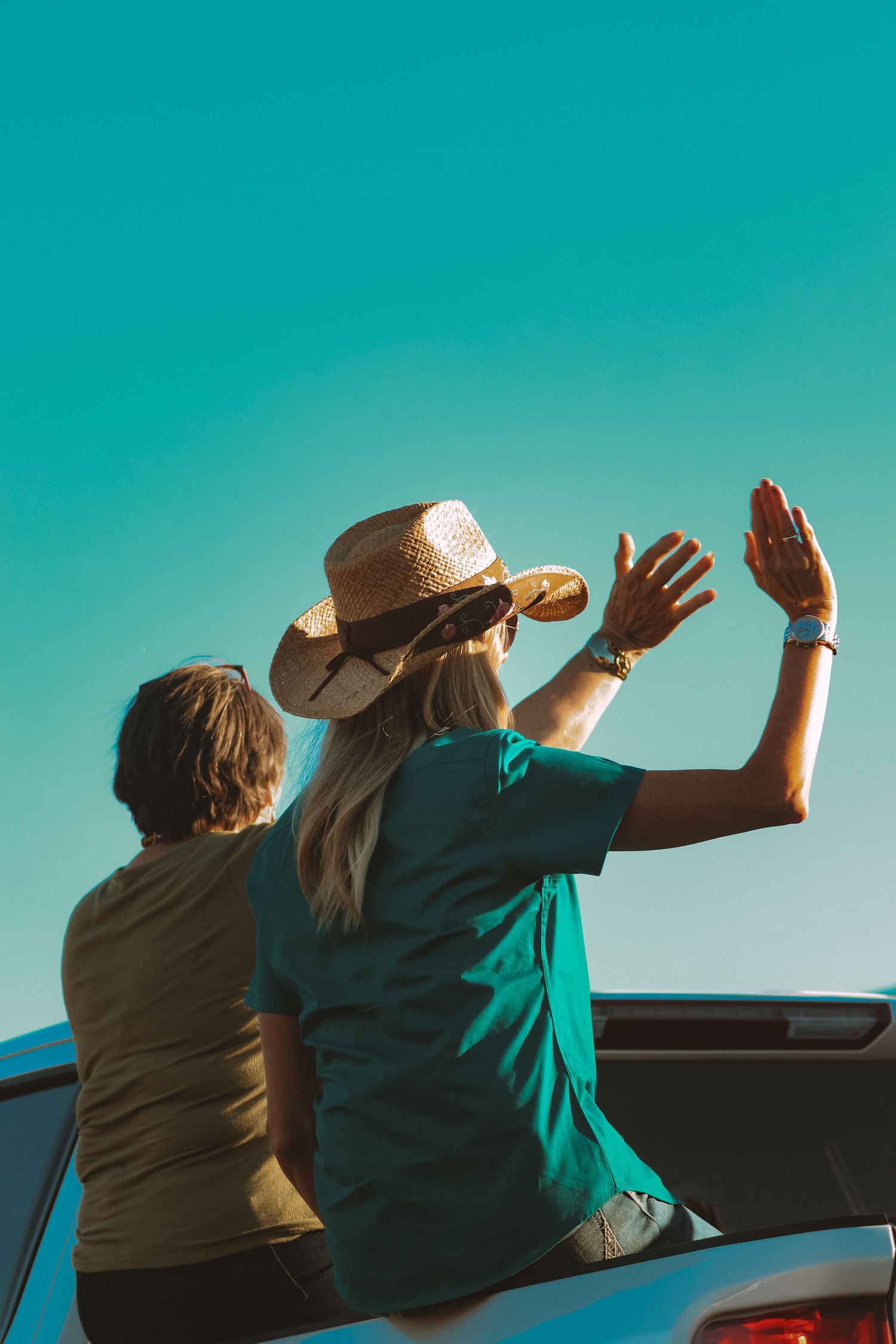 People in cowboy hats wave from car window against turquoise sky.
