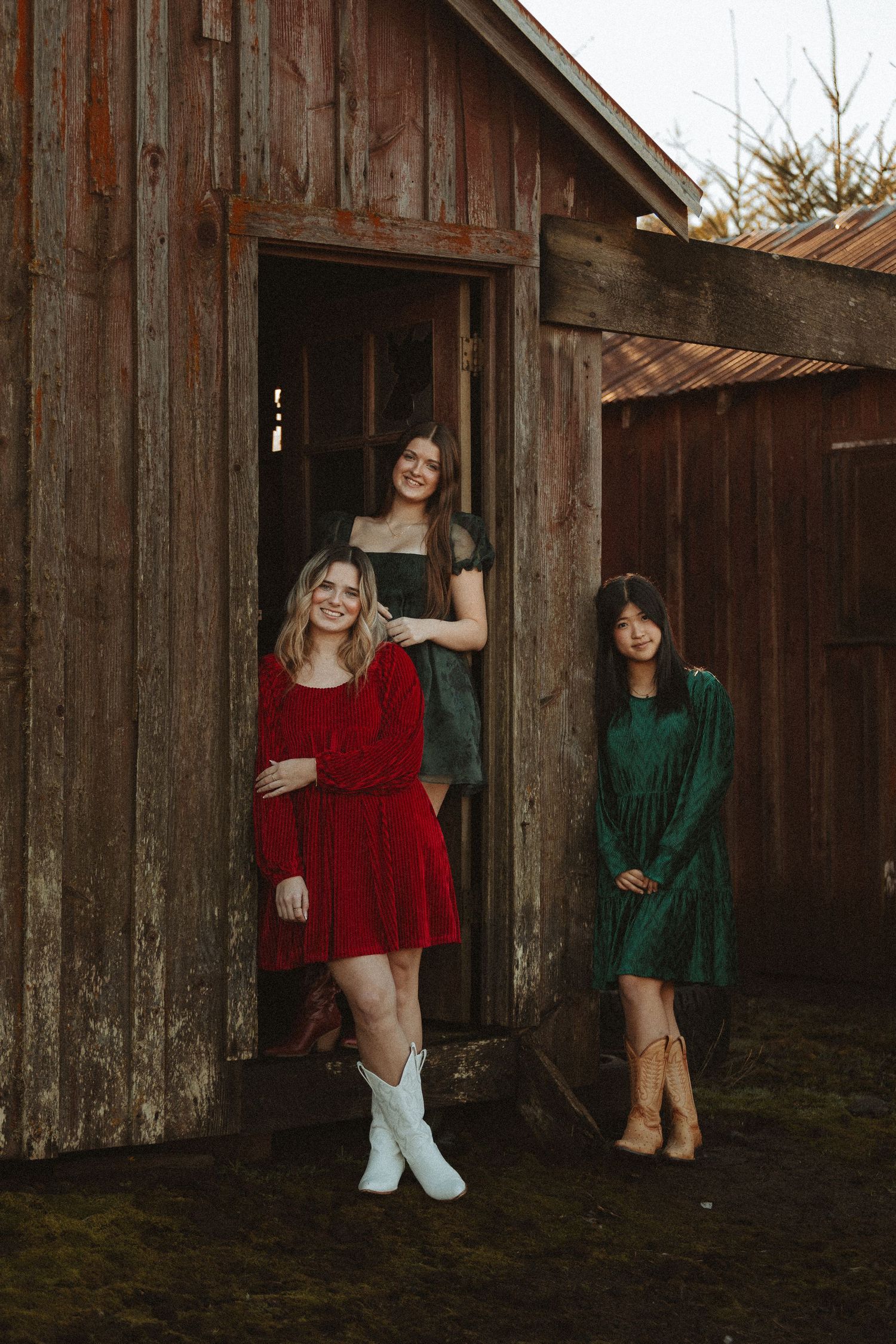 Three people in holiday dresses pose casually by wooden barn entrance.