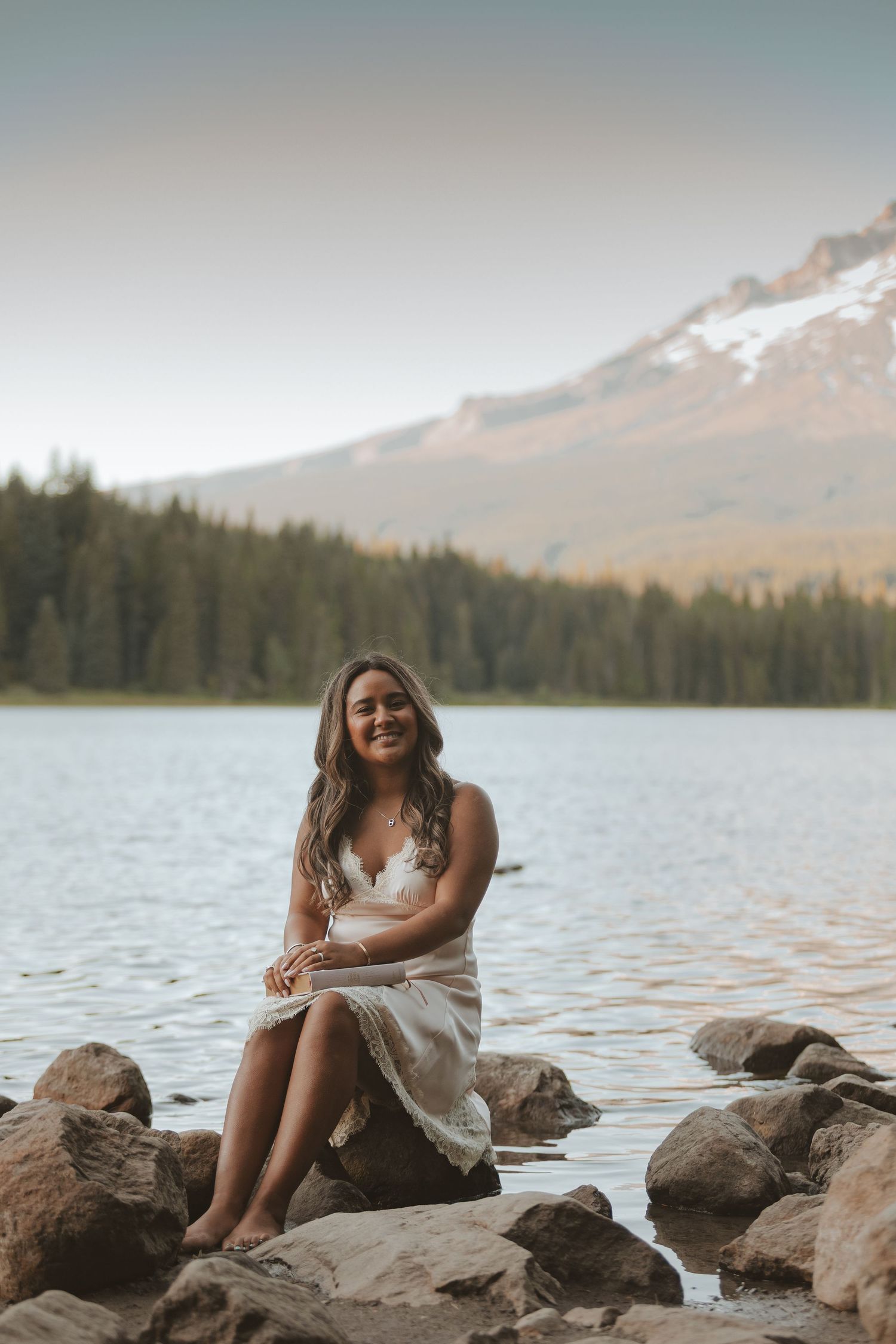 Woman in white dress sits peacefully on rocky shoreline by mountain lake at dusk.