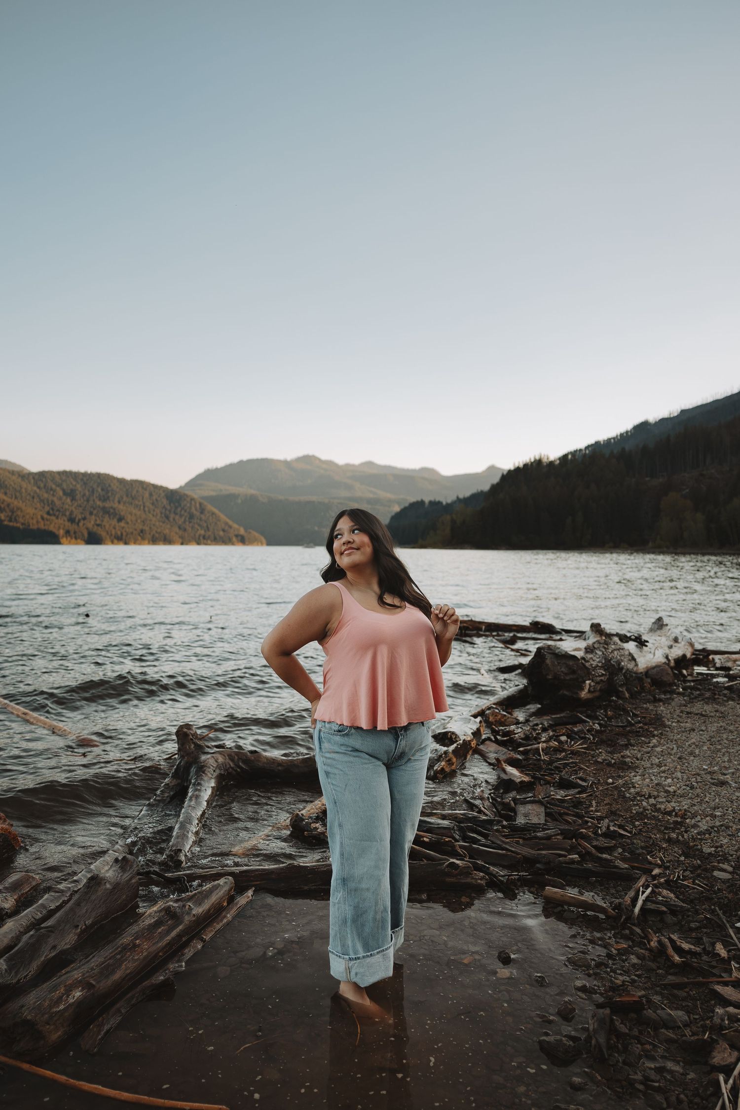 Woman in pink tank top and jeans stands barefoot by a serene mountain lake with driftwood and forested hills.