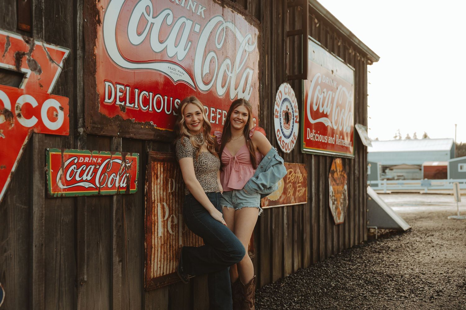Two young women pose against rustic wooden wall covered in vintage Coca-Cola advertising signs outdoors.