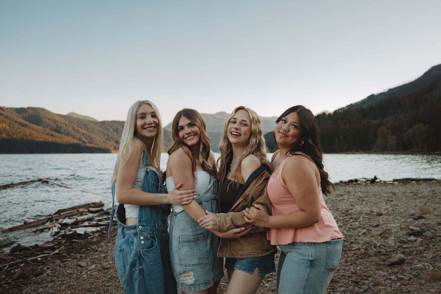 Four smiling women pose together on a rocky beach beside a mountain lake.