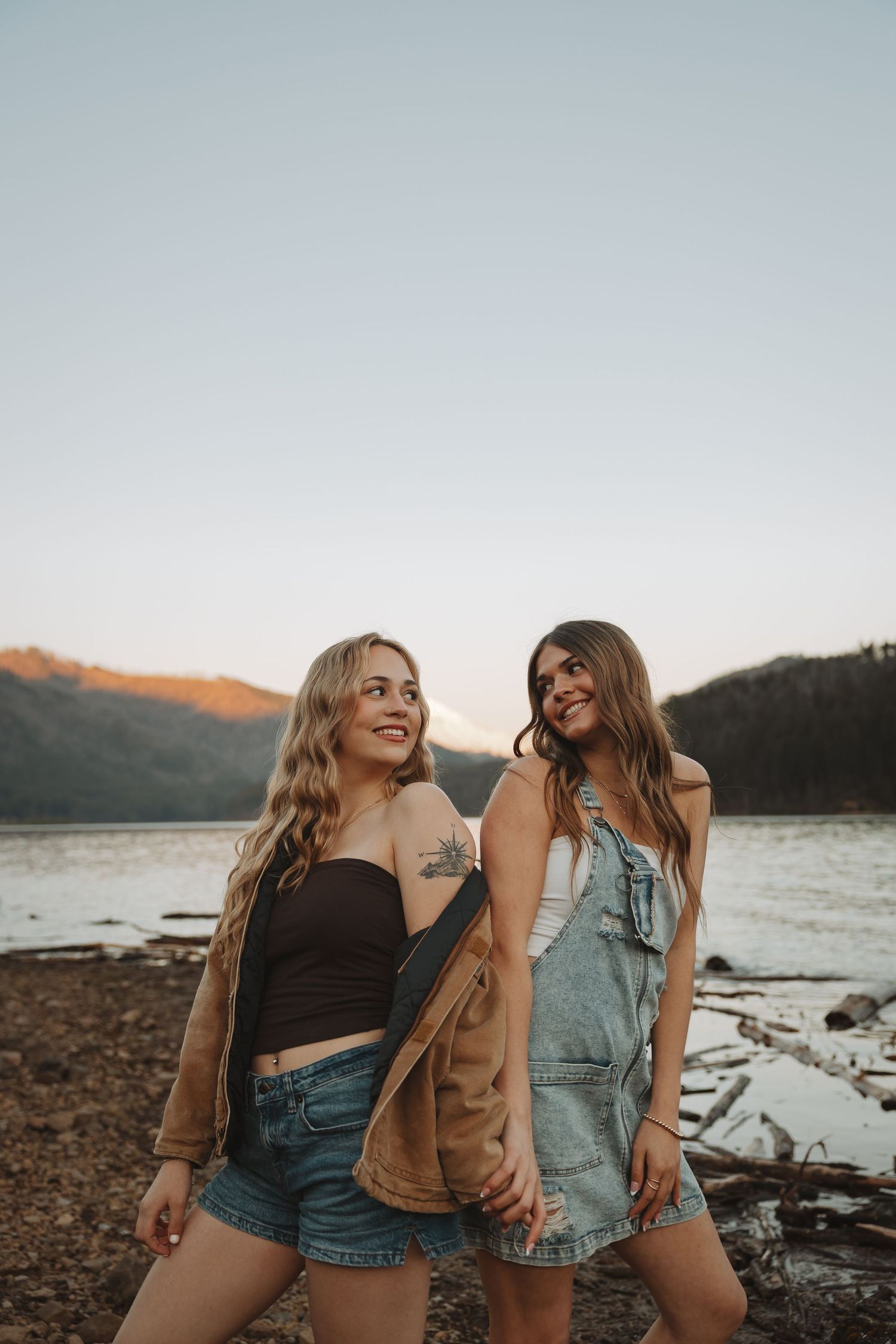 Two young women smiling and holding hands by a mountain lake during golden hour.