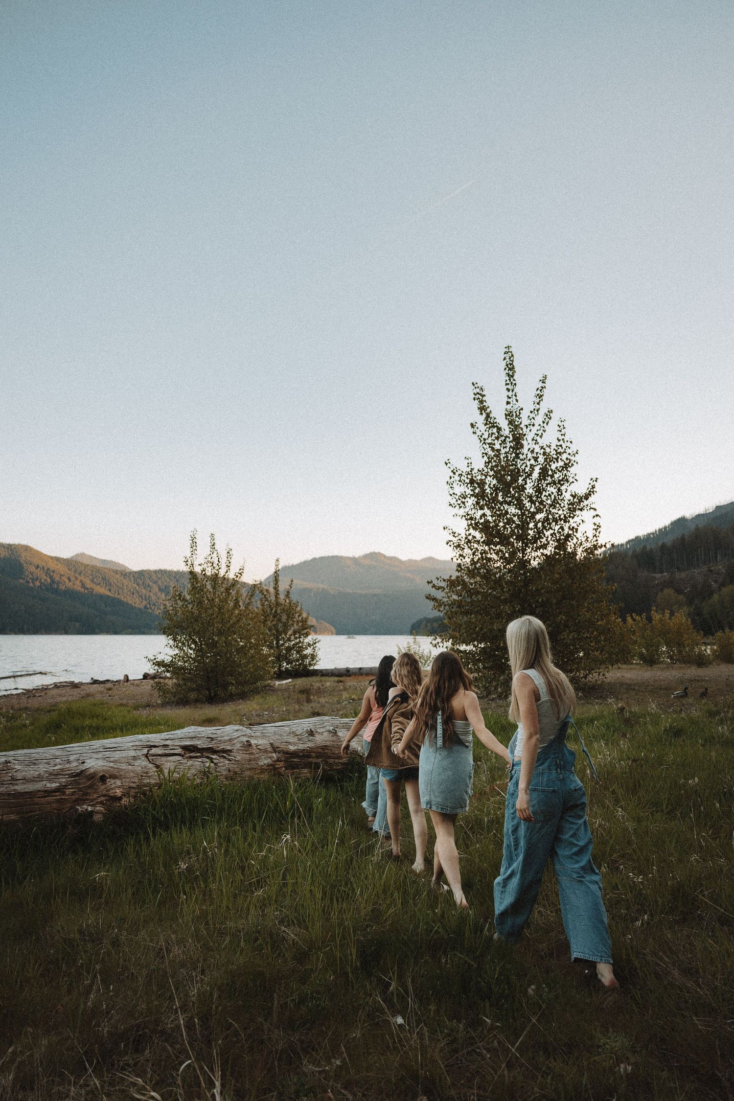 Three young women walking hand in hand through a meadow near a mountain lake.