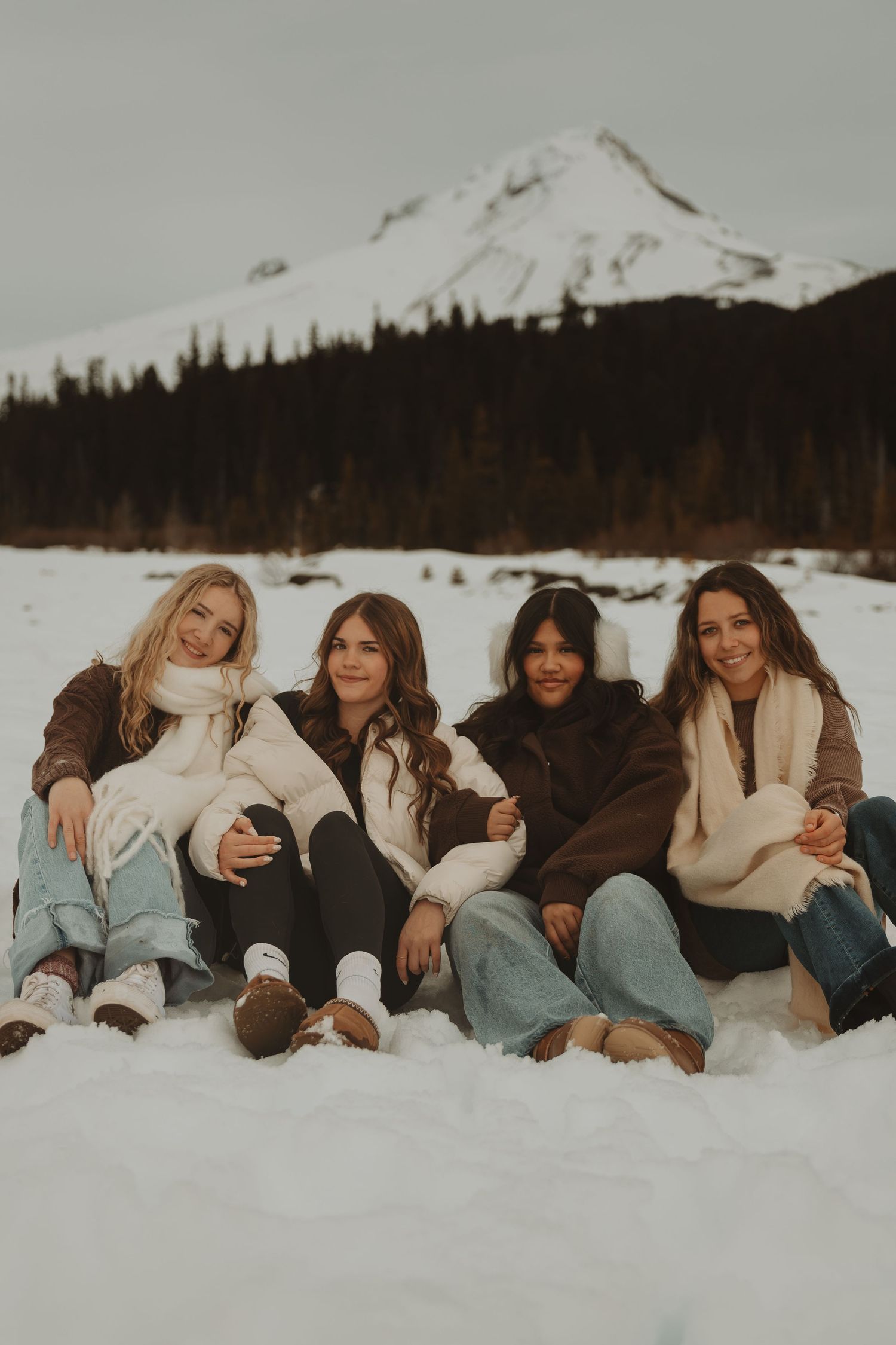 Four women sit together smiling on snow with a snow-capped mountain and pine trees behind them.