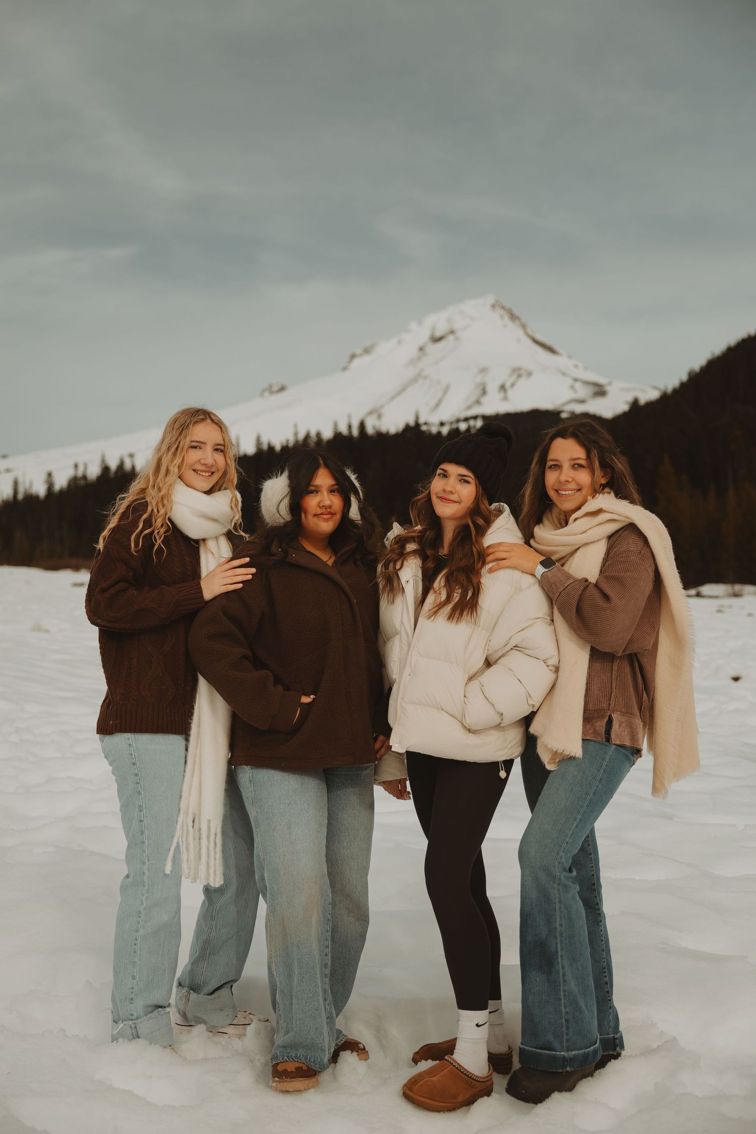 Four women in winter outfits pose together in snowy mountain landscape.