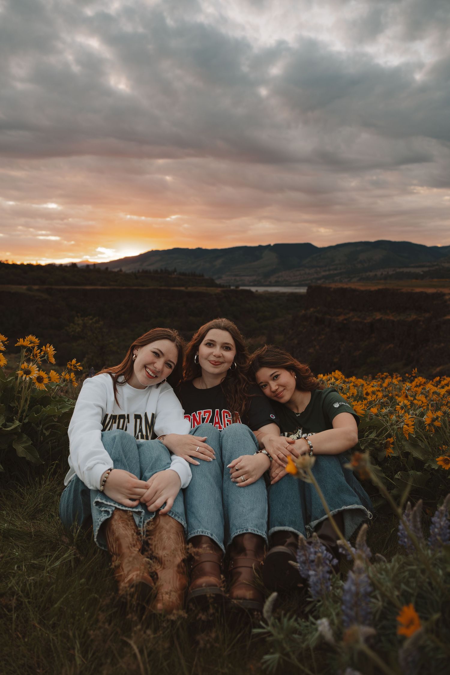 Three smiling girls sitting together in a wildflower field during a colorful sunset with mountain views.