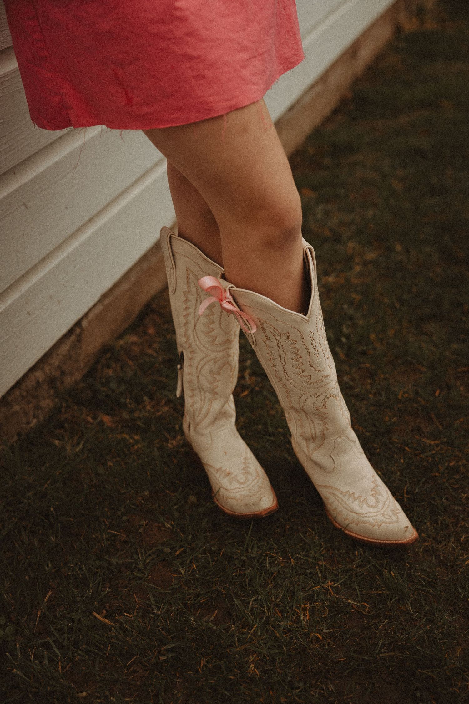 White cowboy boots with decorative stitching shown against dark ground beneath a red dress hem.