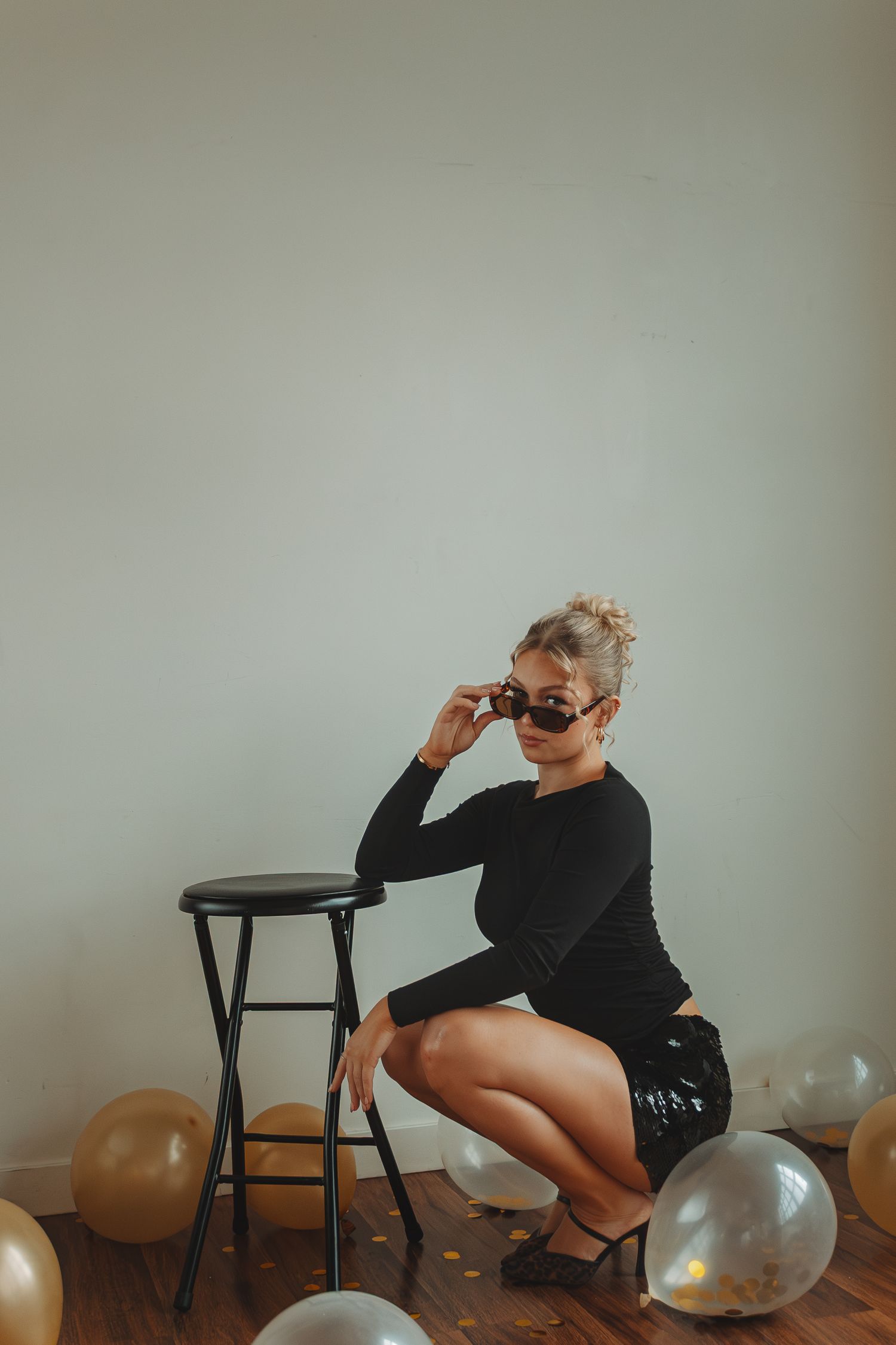 Fashion model in black poses artistically with balloons on hardwood floor against neutral wall.