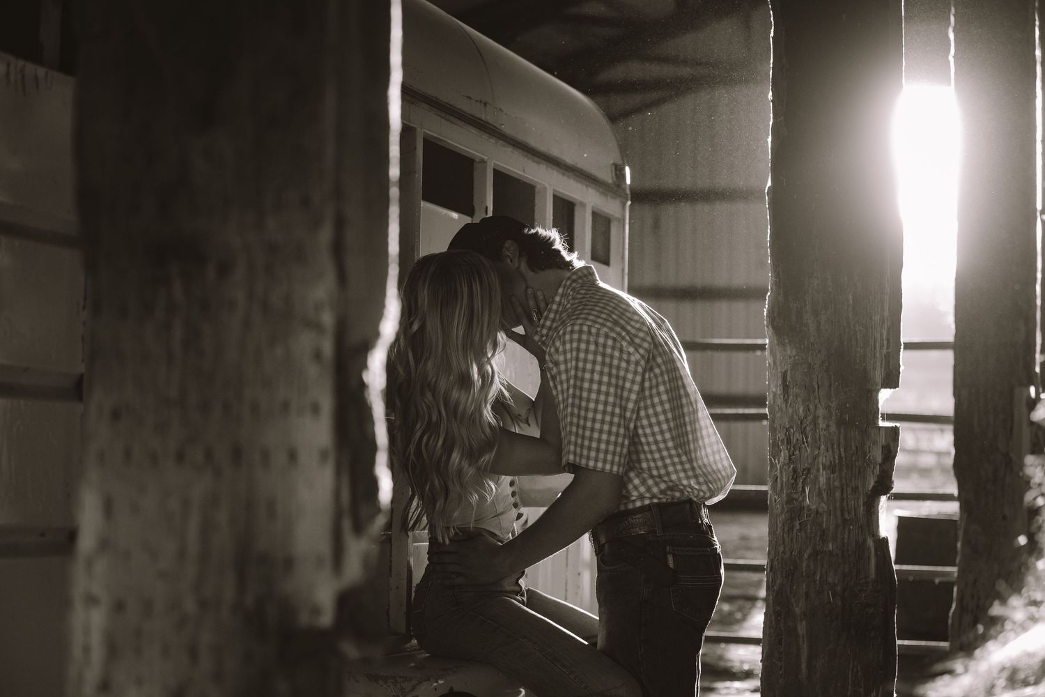 Black and white photo of couple sharing a kiss near a vintage train car in warm backlight.