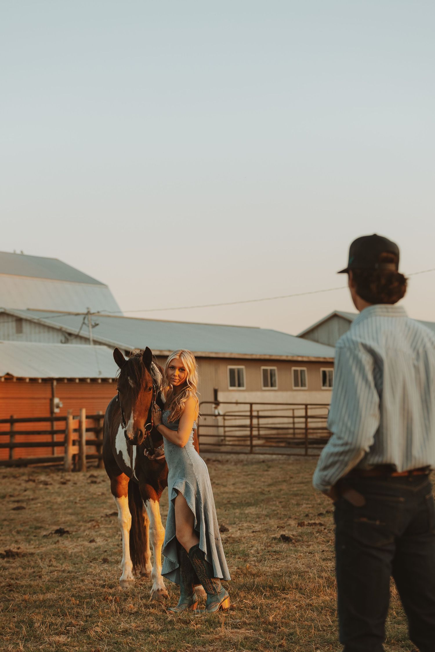 Blonde woman in blue dress stands with horse as man watches at sunset farm.