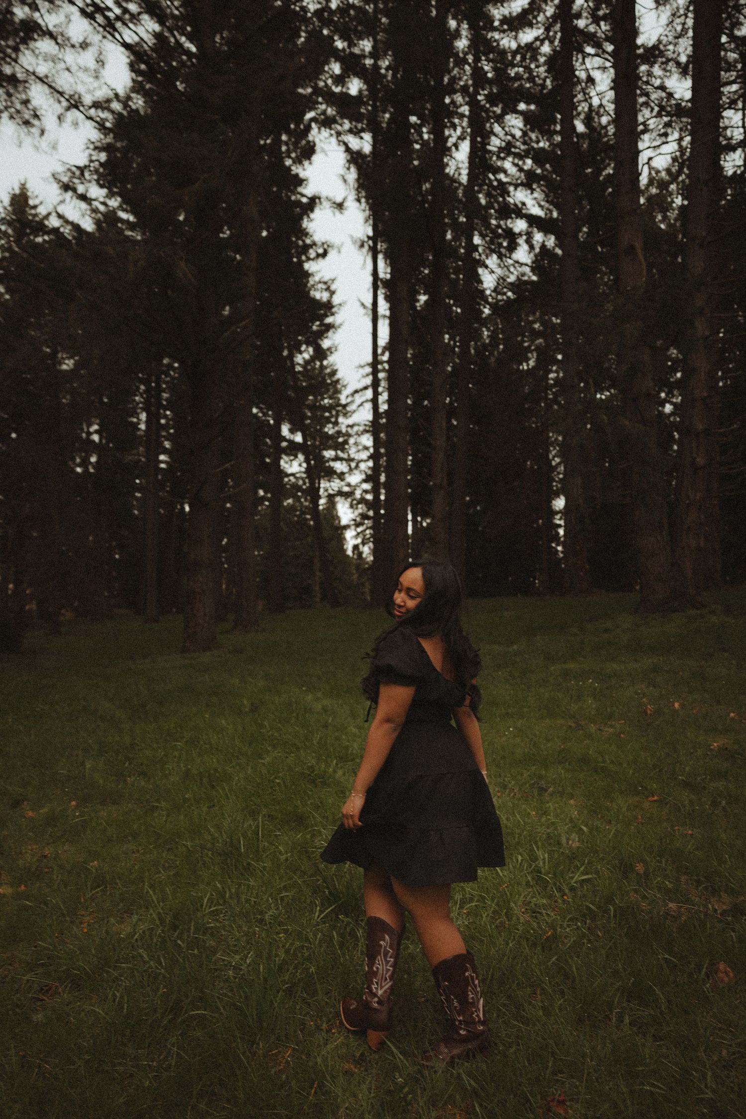 A person in dark clothing crouches in a forest clearing at dusk among tall evergreen trees.
