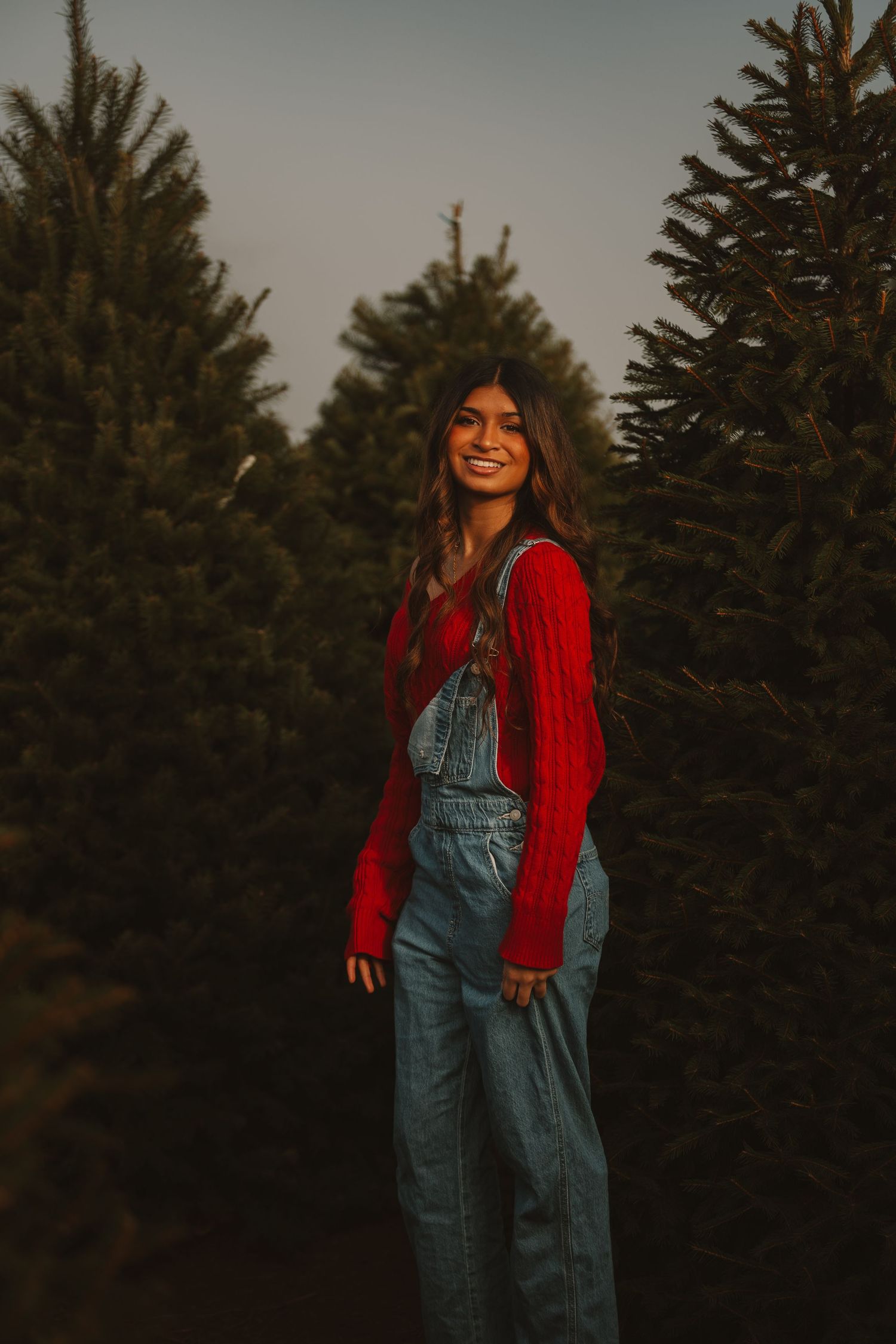 Person in denim overalls and red sweater poses joyfully among evergreen trees at sunset.