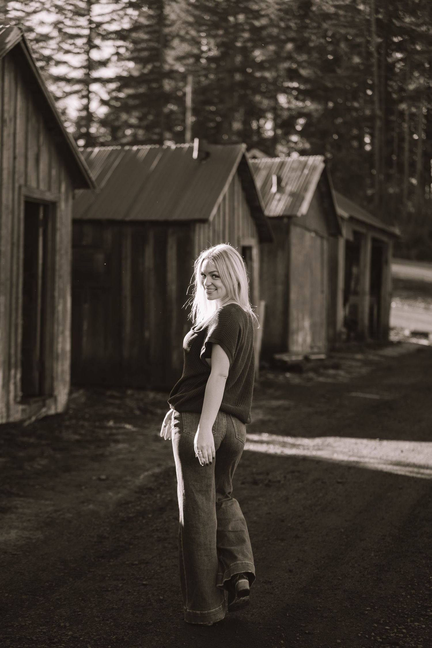 Black and white portrait of person standing near rustic wooden buildings.