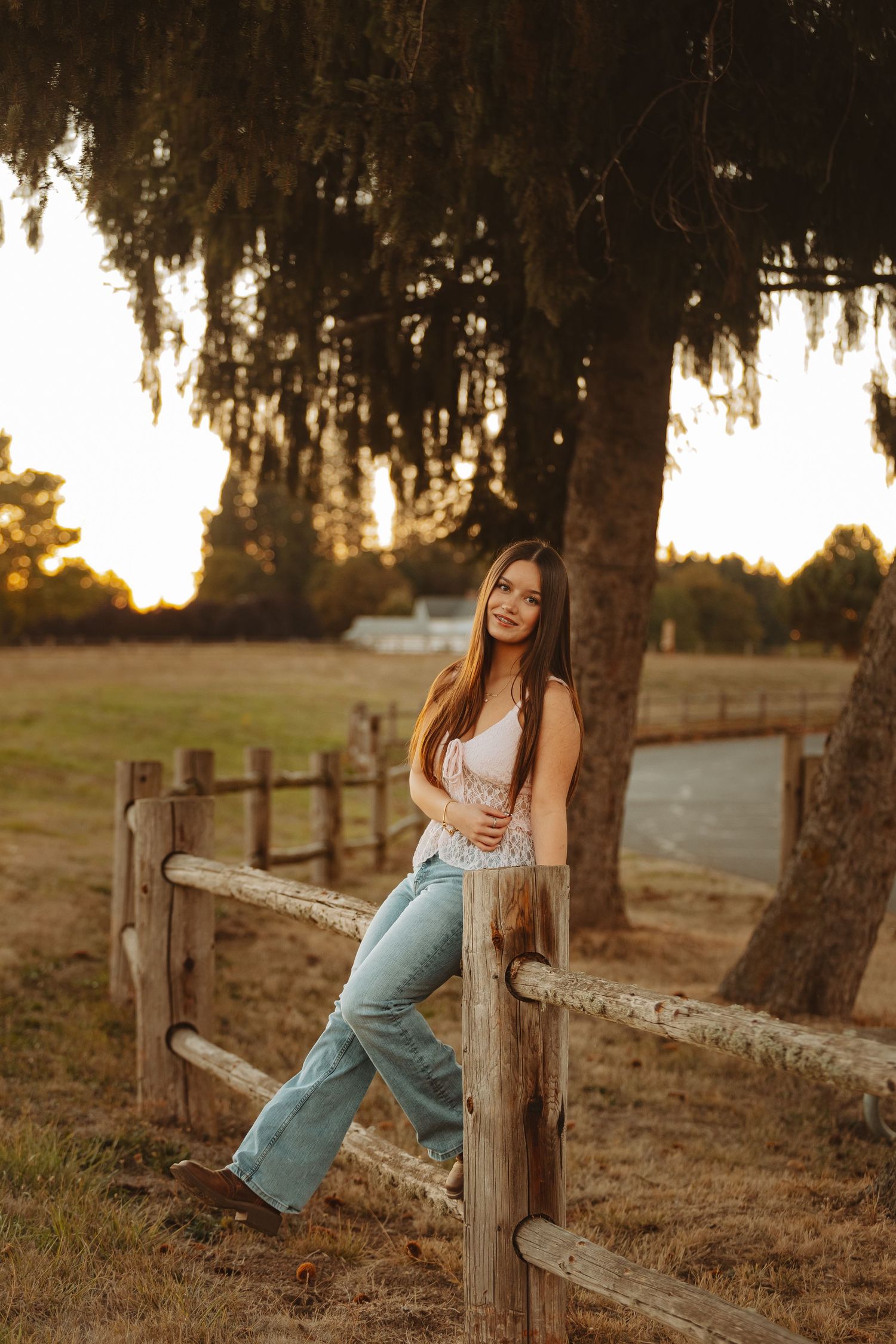 A person in jeans sits casually on a wooden fence at sunset with trees silhouetted in the background.