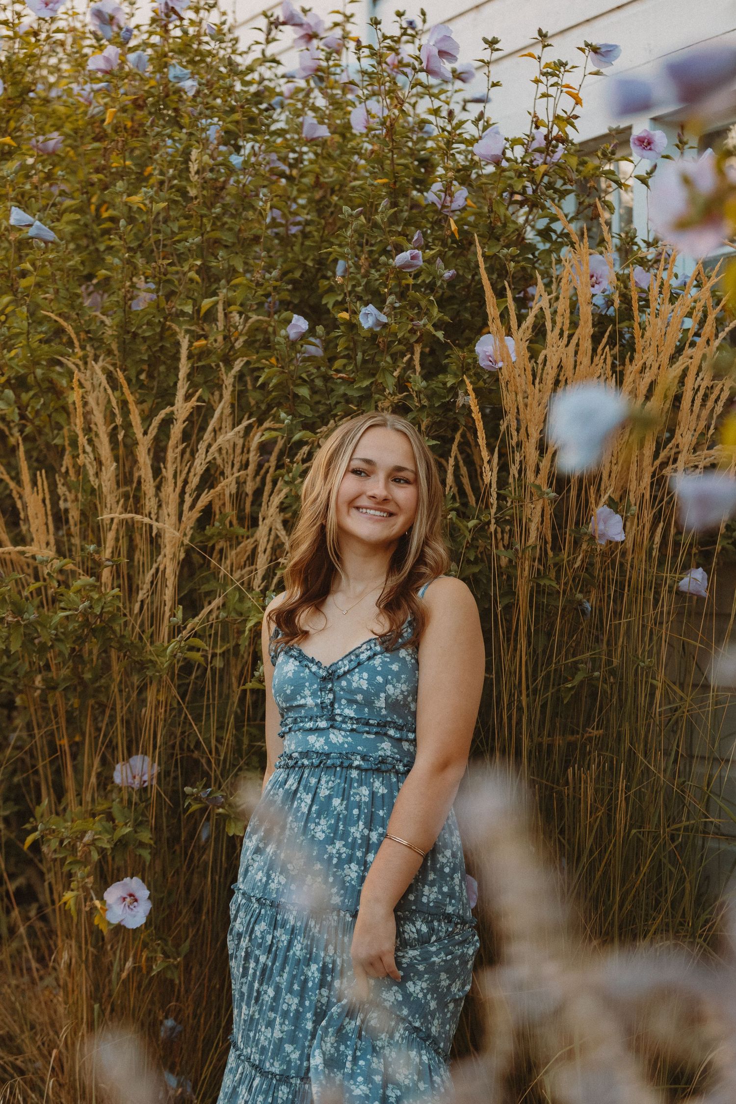 A person in a blue floral dress poses in front of tall dried grasses and autumn foliage during a golden hour photoshoot.