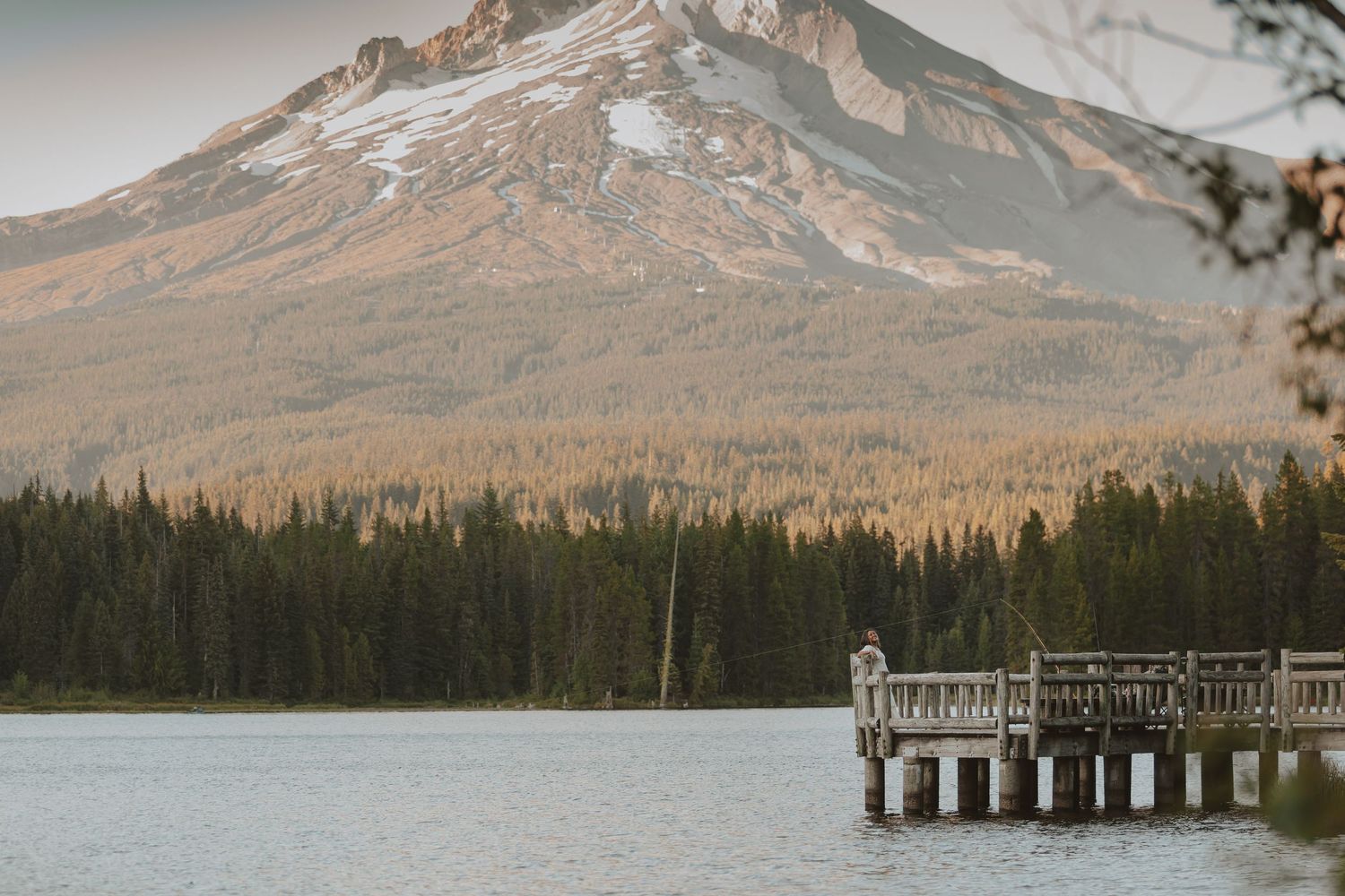 Snow-capped Mount Hood towers over a serene lake with a wooden dock extending into calm waters at sunset.