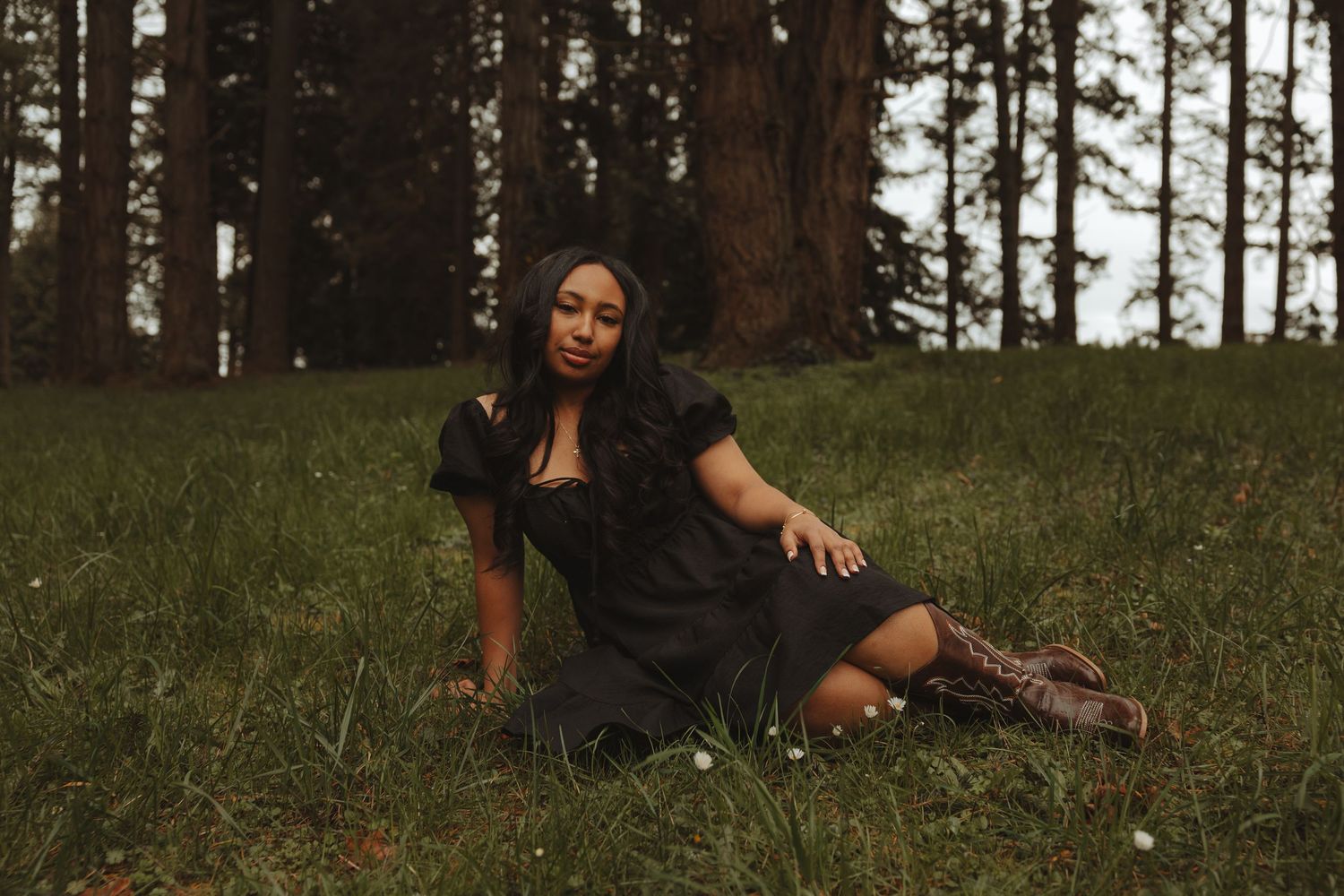A person in black clothing poses gracefully in a grassy field with tall pine trees in the background.